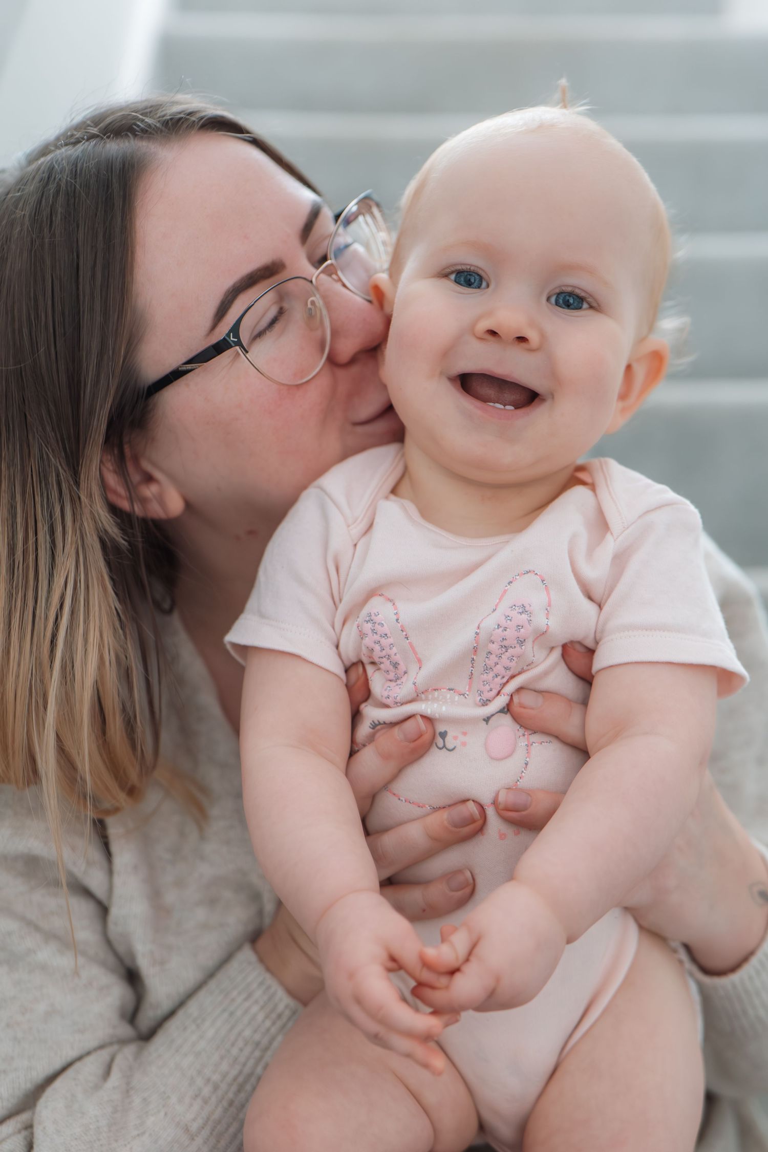 Mother with glasses kissing her laughing baby girl wearing a pink bunny onesie on the cheek.