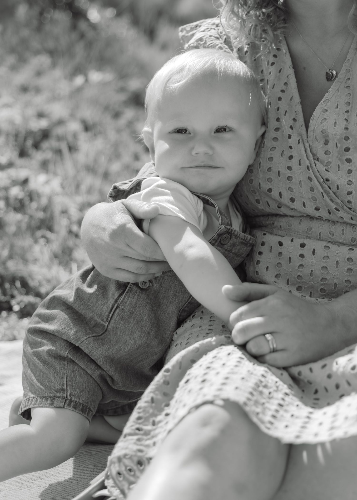 Black and white photo of a baby sitting on a woman's lap outdoors, wearing denim overalls.