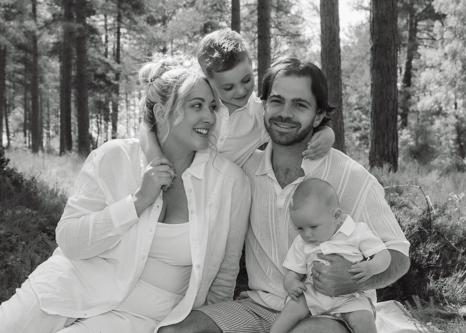 Black and white family portrait of parents with two young children smiling outdoors in a forest setting.