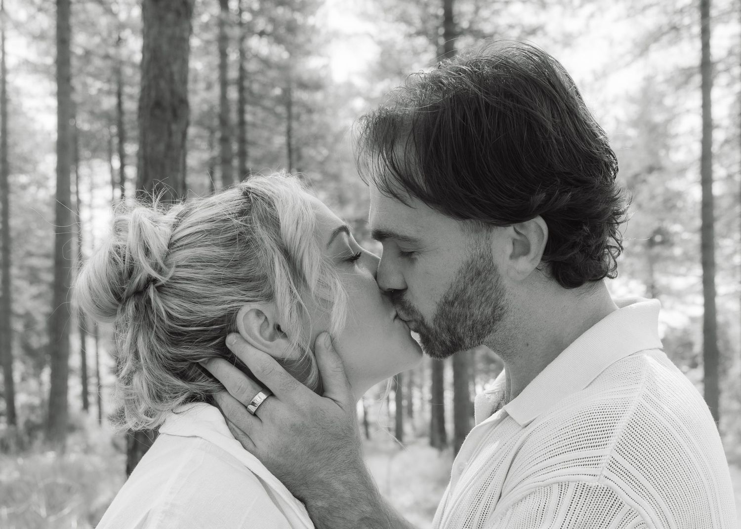 Black and white photo of a couple sharing a romantic kiss outdoors among tall trees.