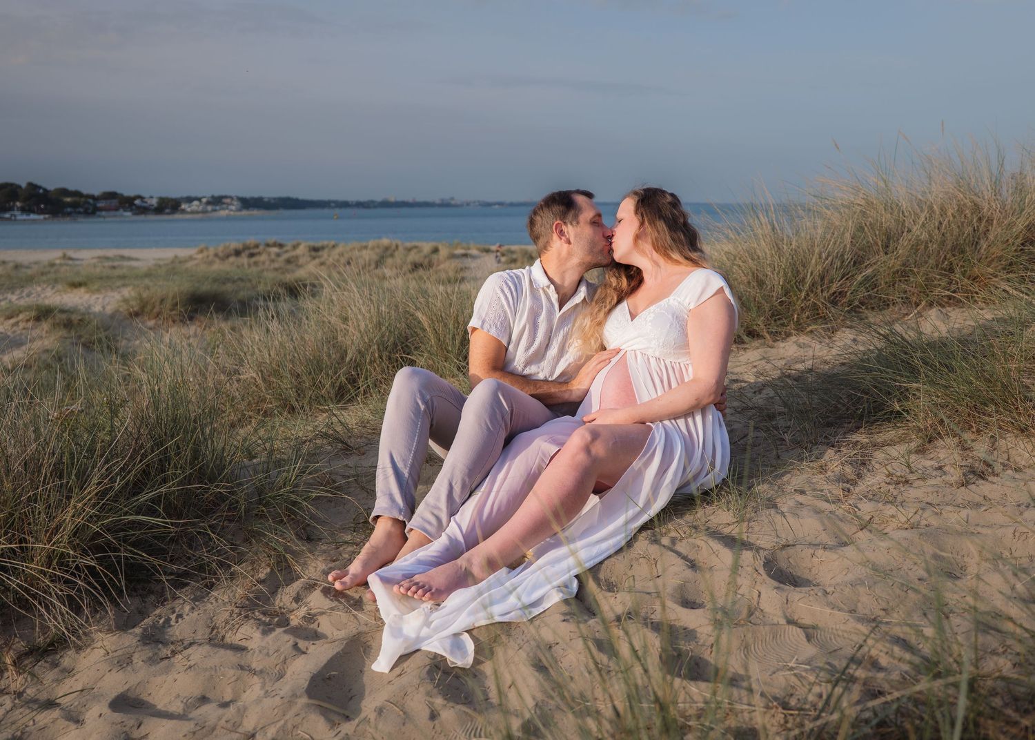 Couple sharing a kiss on sandy beach dunes, woman in white maternity dress, golden hour light, seaside backdrop.
