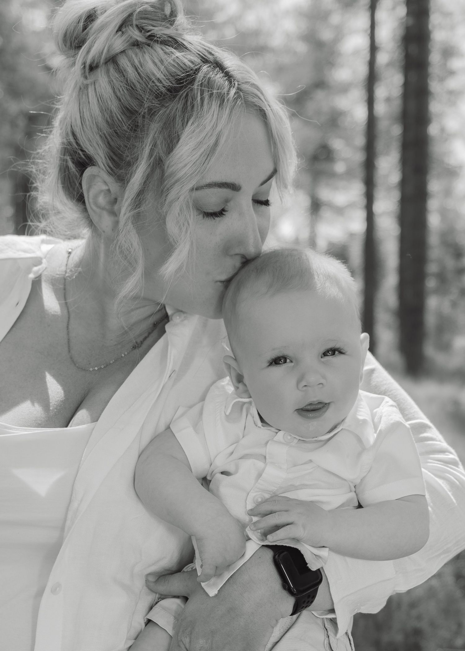 Black and white photo of a young blonde mother kissing her baby boy outdoors among trees.