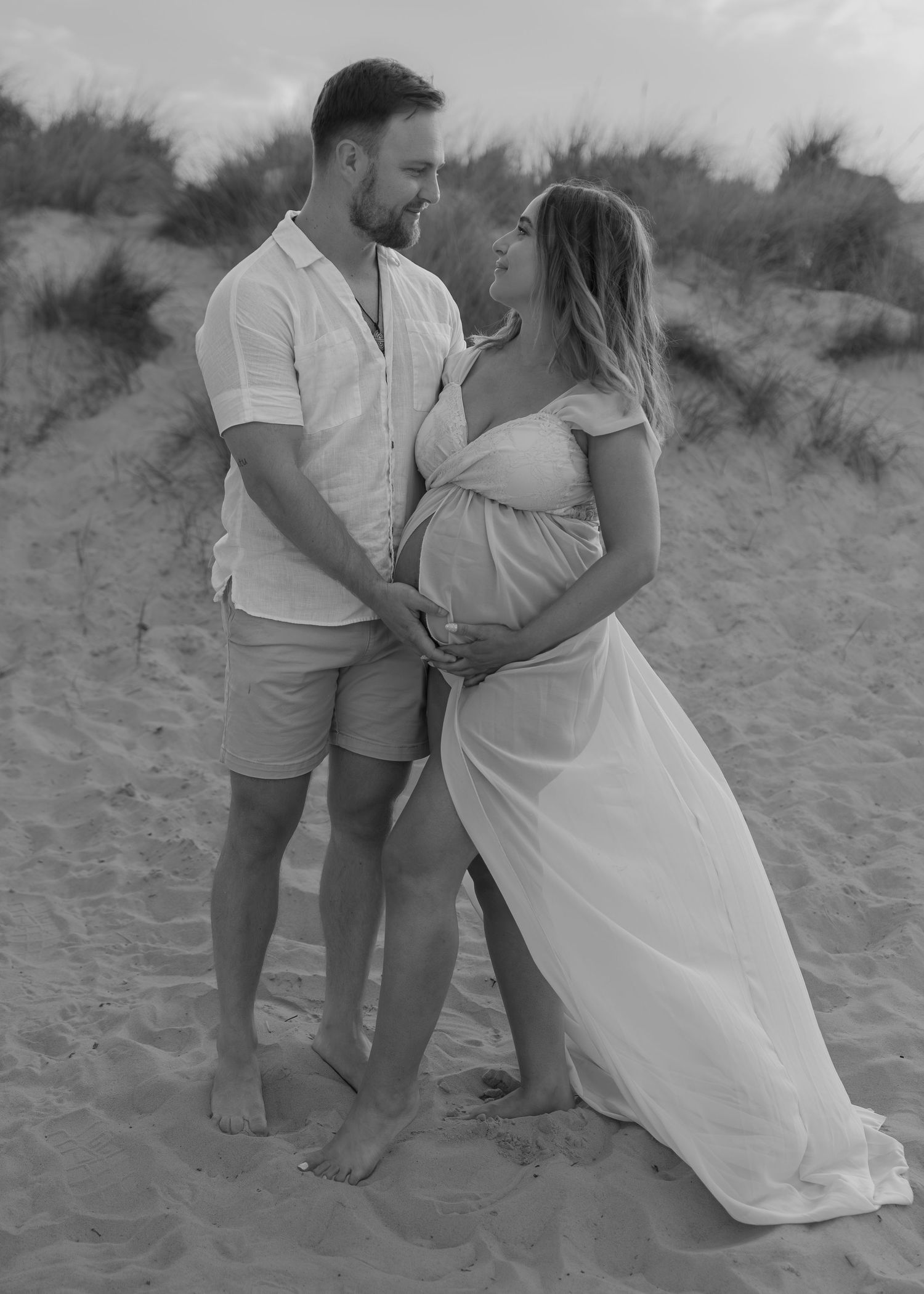 Black and white maternity photo of a couple holding baby bump on sandy beach dunes, woman in flowing white dress.