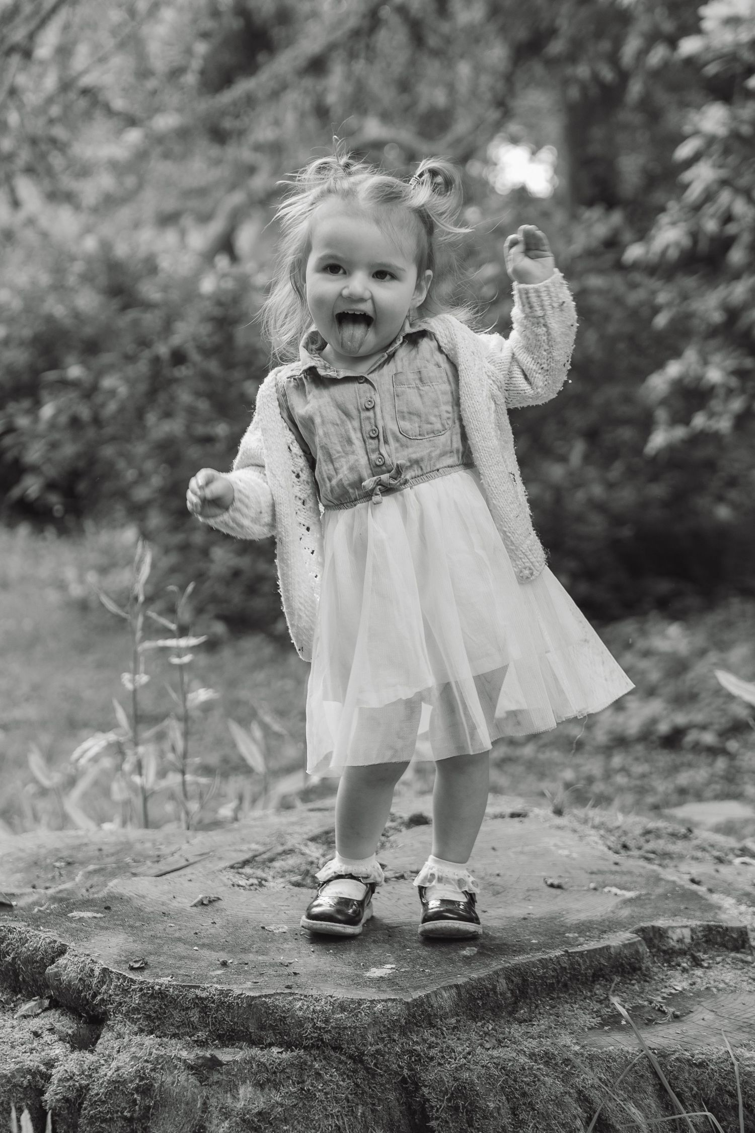 Black and white photo of a playful toddler girl sticking out her tongue while standing on a stone outdoors.