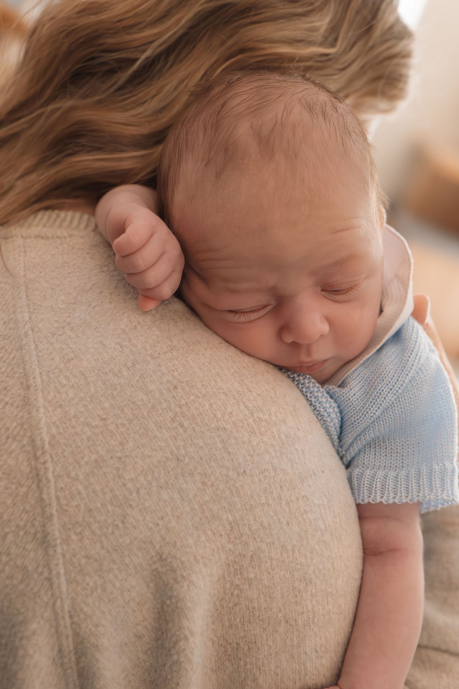 Newborn baby sleeping peacefully on mother's shoulder wearing a light blue knit outfit.