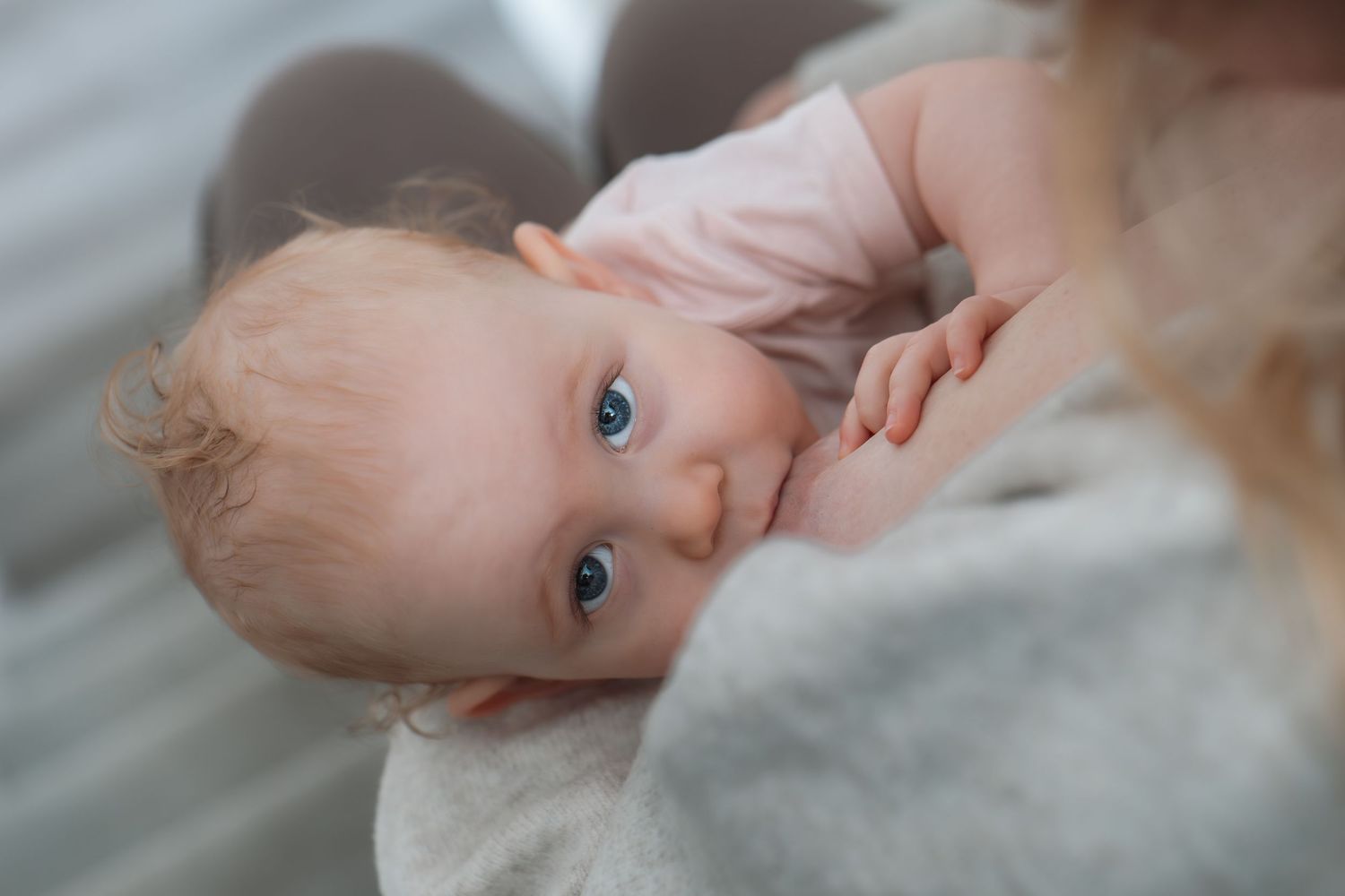 Close-up of a blue-eyed baby breastfeeding while gazing up with a soft blanket nearby.