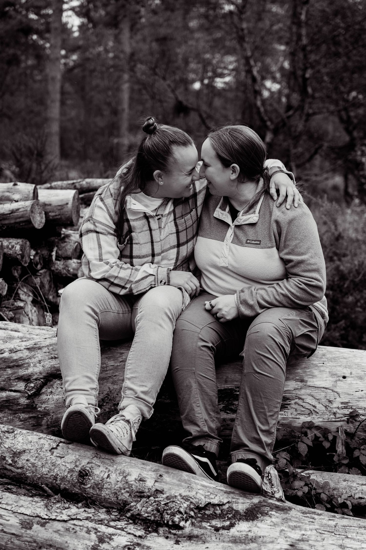 Two women sitting on logs in a forest, sharing an intimate moment touching foreheads, black and white photo.