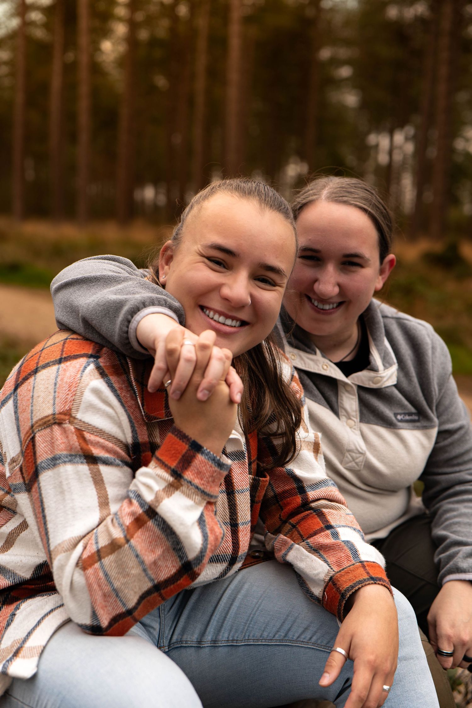 Two smiling women sitting together in a forest, one resting her head on the others shoulder.