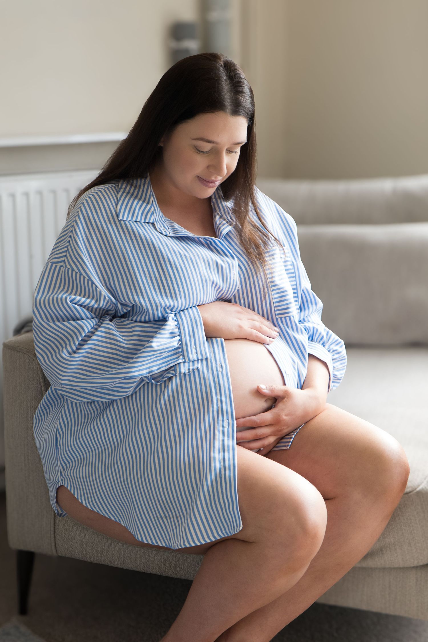 Pregnant woman in blue striped shirt sitting on sofa gently cradling her baby bump indoors.