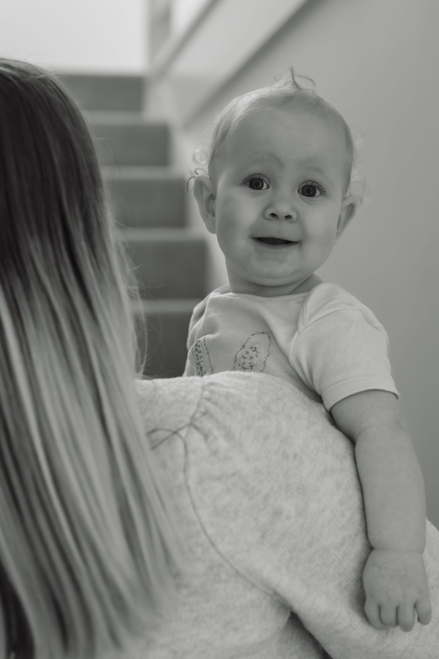 Black and white photo of a happy baby being held by a woman, smiling at the camera near a staircase.
