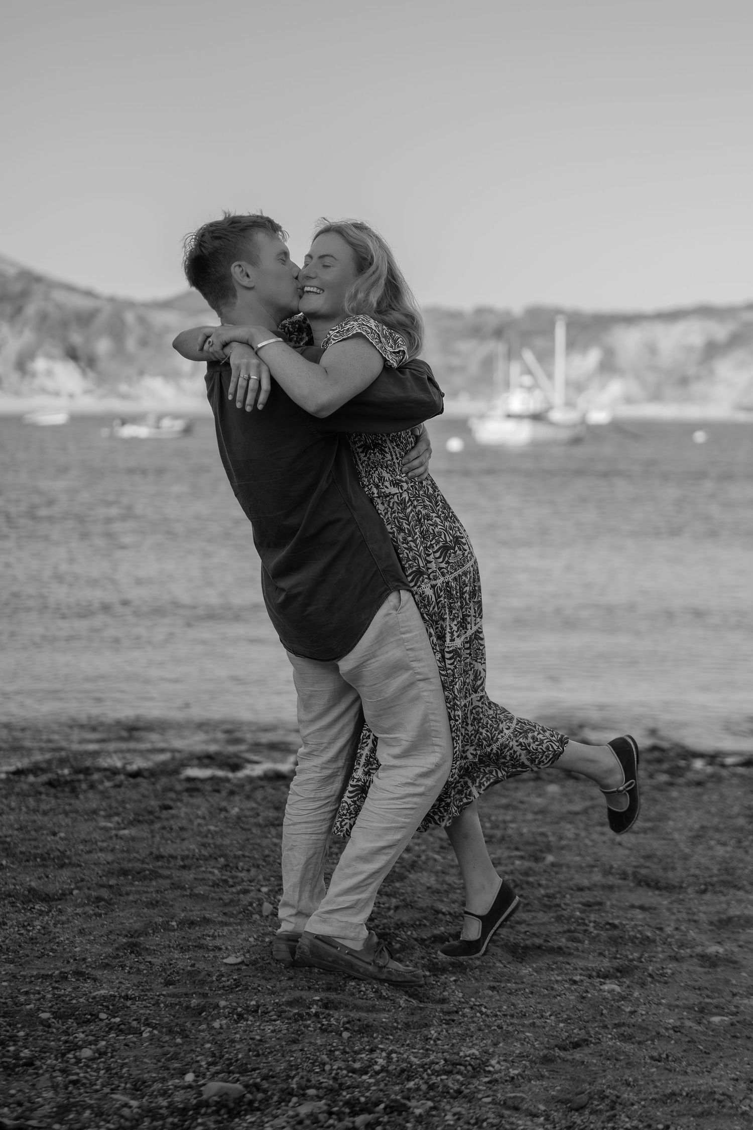 Black and white photo of a couple embracing and laughing on a rocky beach with sailboats in the background.
