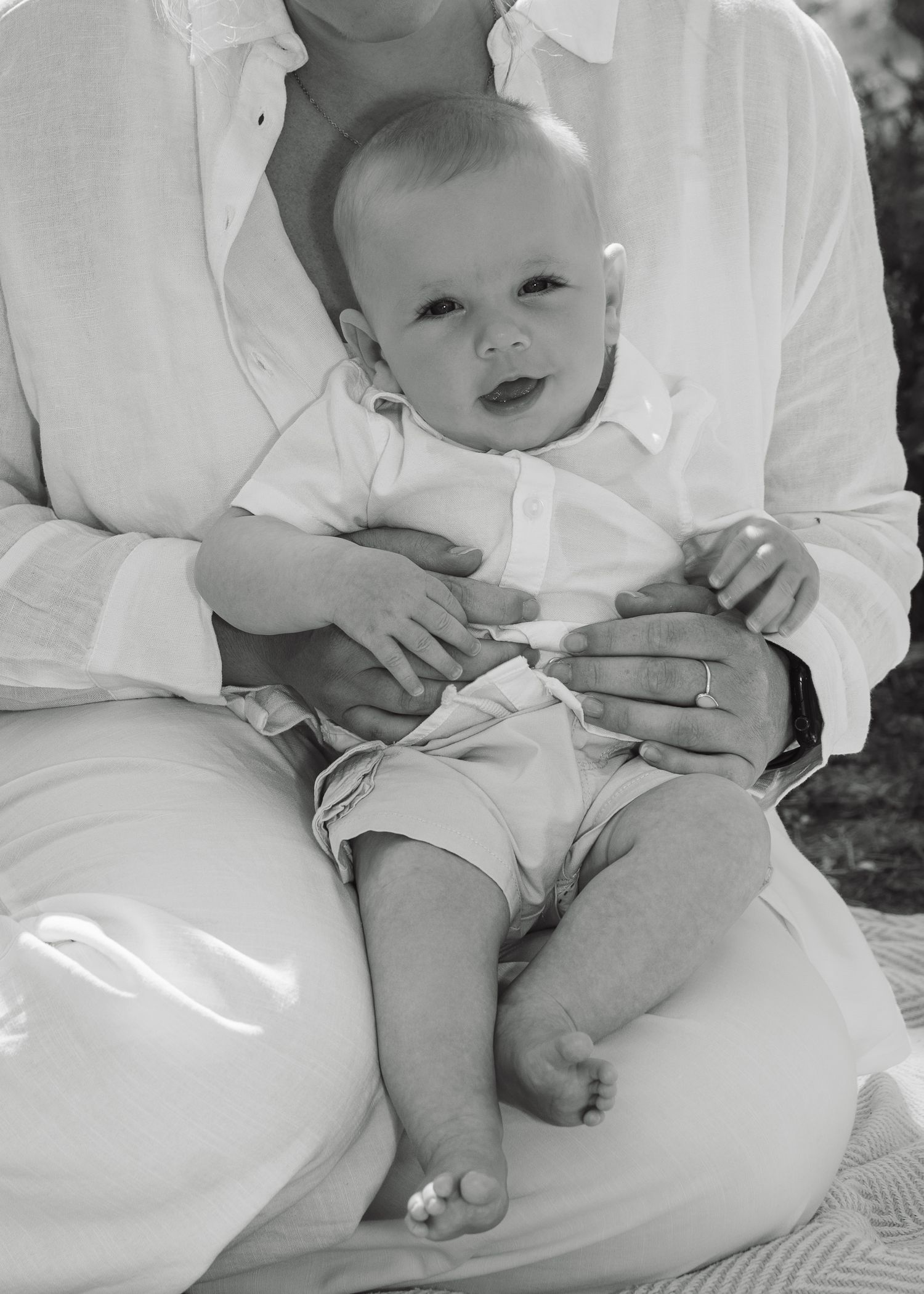 Black and white photo of a smiling baby sitting on a parent's lap, both dressed in white clothing outdoors.