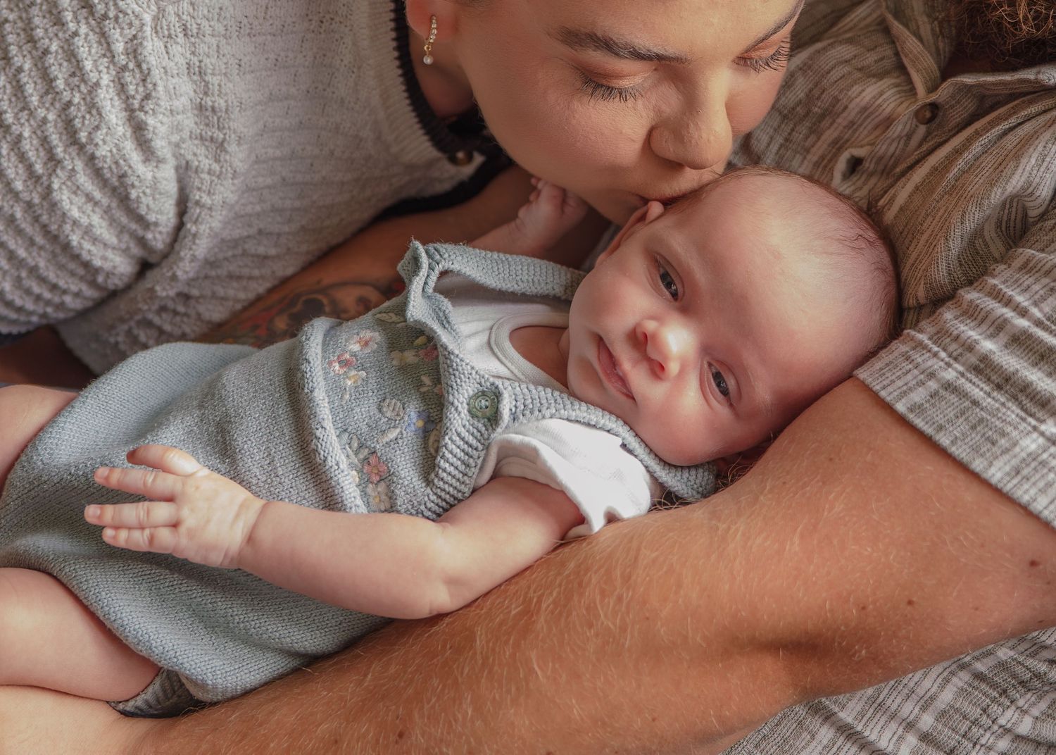 A mother tenderly kisses her newborn baby while the infant rests on a parent's chest in a cozy setting.
