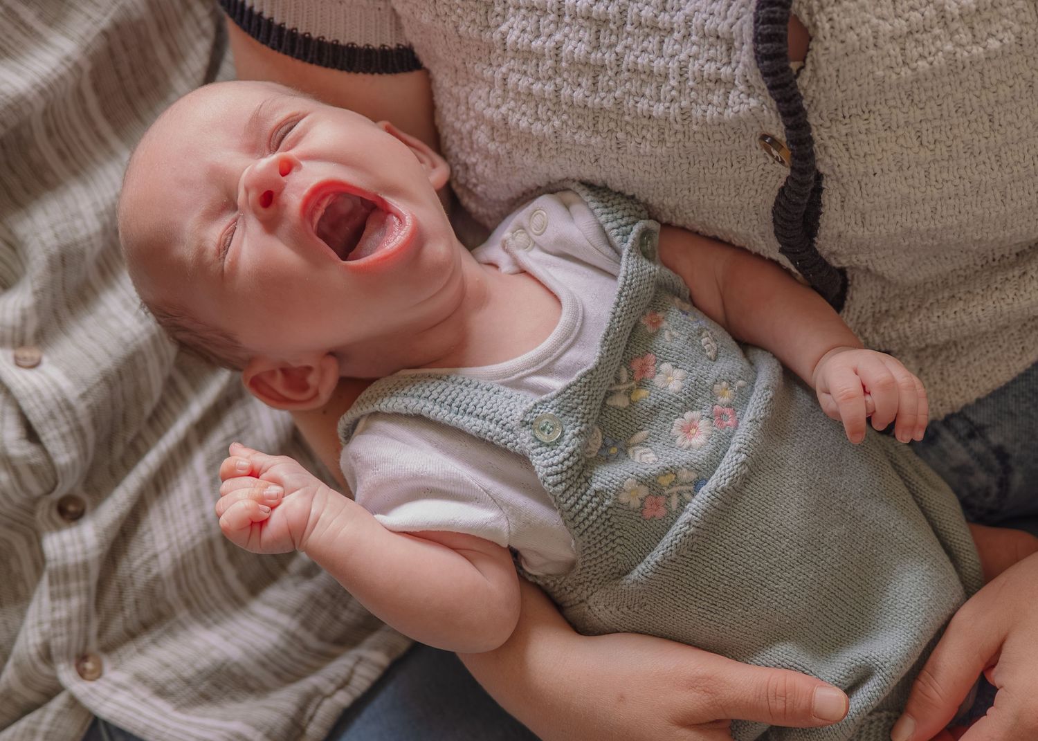 A crying baby in a green floral outfit being held by two adults, showing distress with clenched fist.