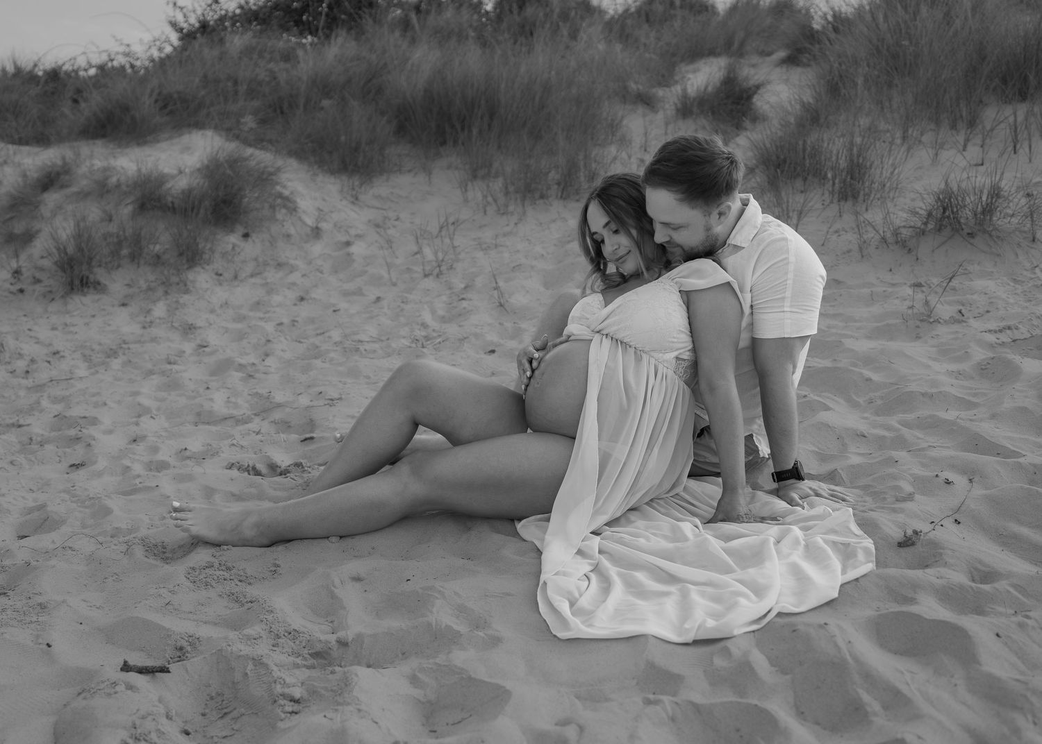 Black and white maternity beach photo of a couple sitting on sand dunes, woman in flowing white dress cradling baby bump.