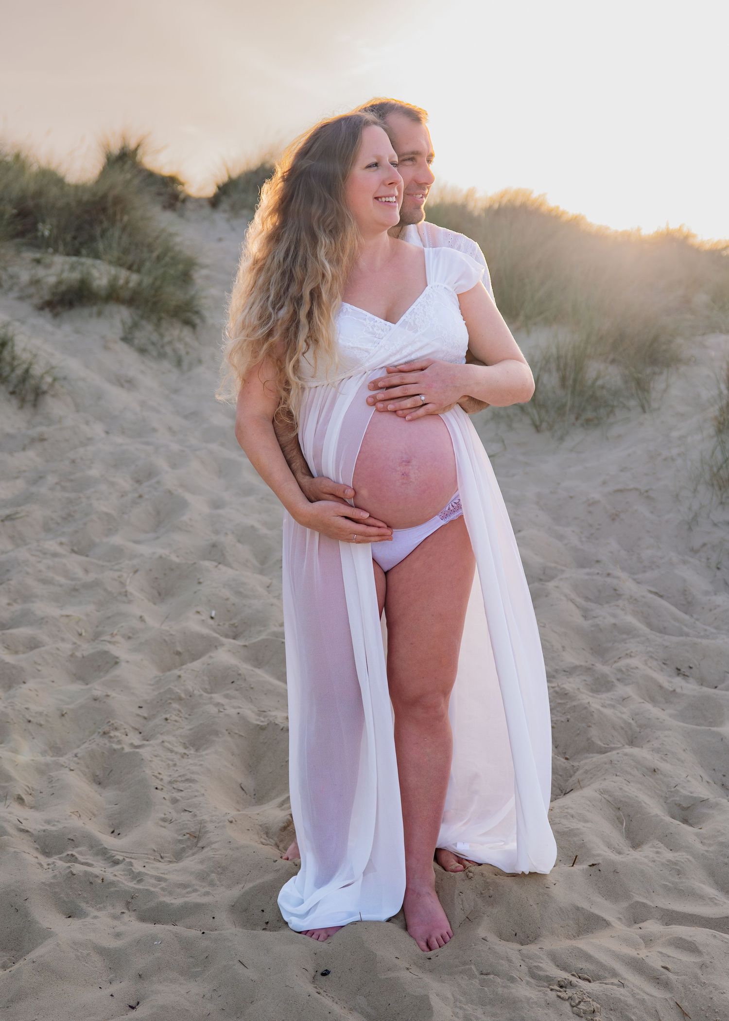 Pregnant couple embracing on sandy beach at golden sunset, woman in white sheer maternity gown cradling baby bump.