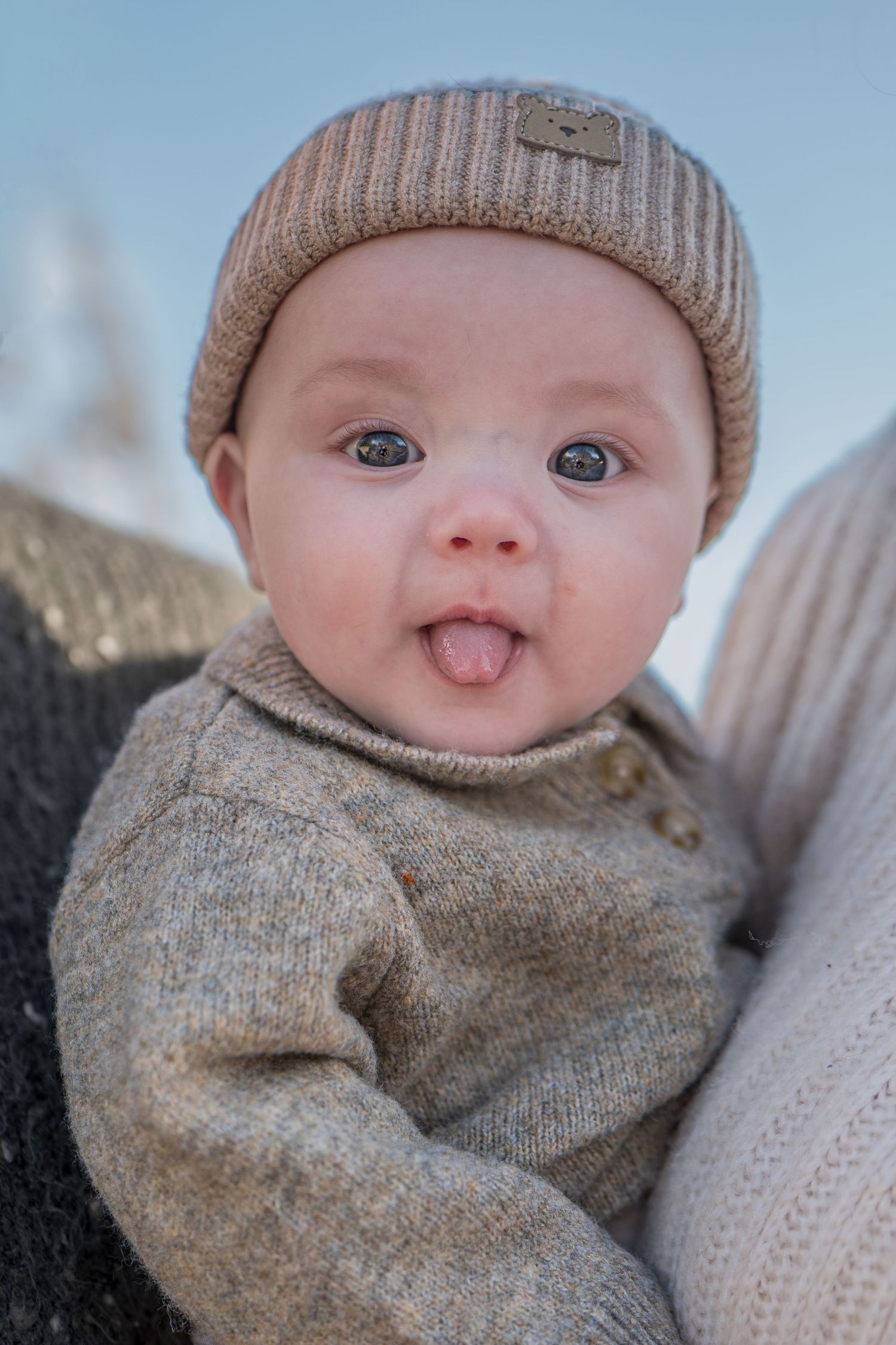 Adorable baby wearing a knit beanie hat sticking tongue out while being held outdoors.
