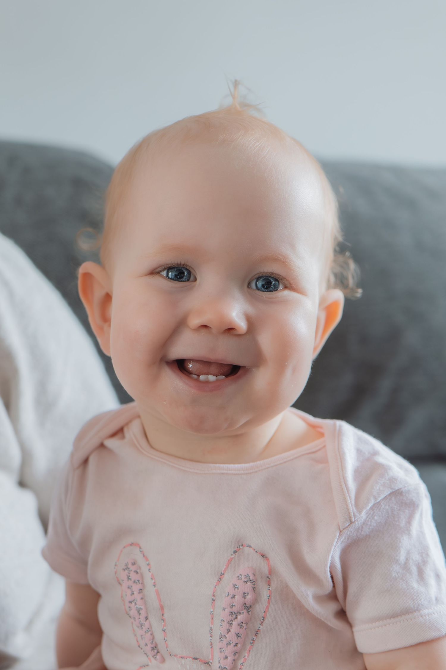 Smiling baby girl with blue eyes wearing a pink bunny onesie sitting in front of a gray sofa.