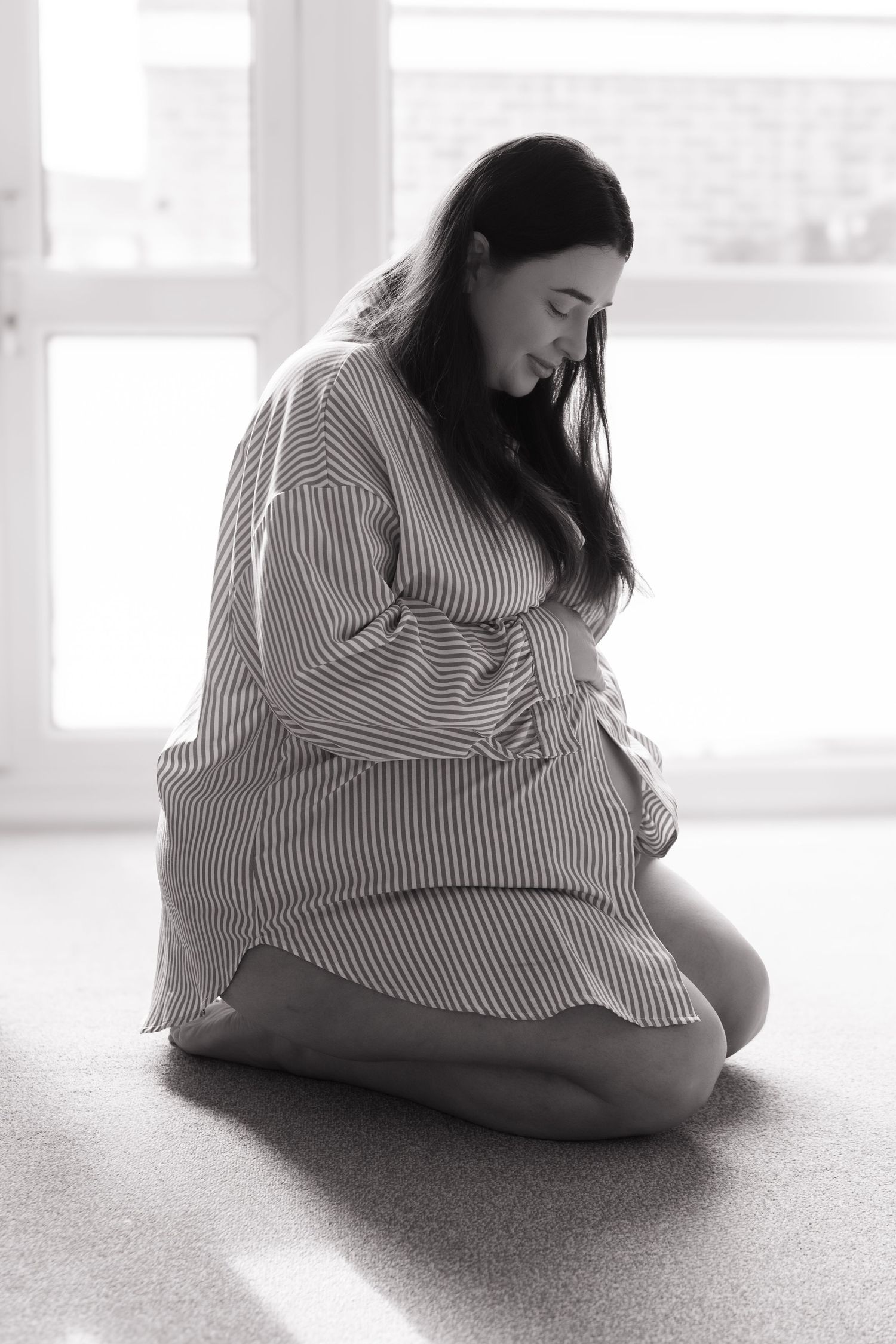 Black and white photo of pregnant woman in striped shirt kneeling on floor near bright window.