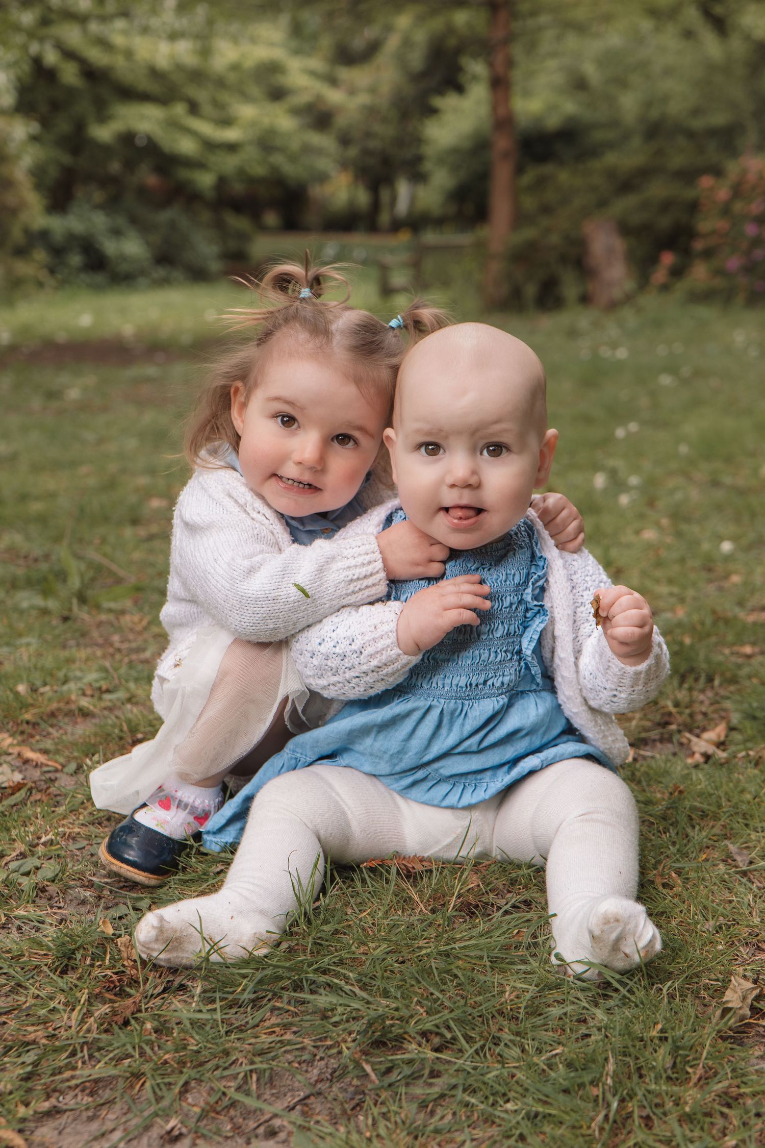 Two young sisters pose together on grass in a park, the older girl hugging the baby from behind warmly.