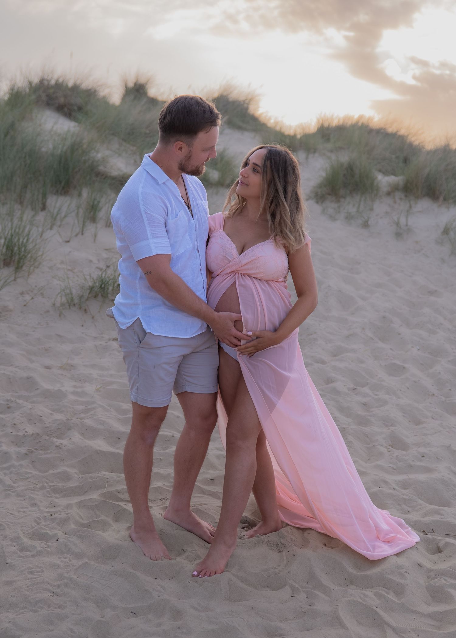 Couple in maternity photoshoot on sandy beach at sunset, woman in pink flowing gown cradling baby bump.