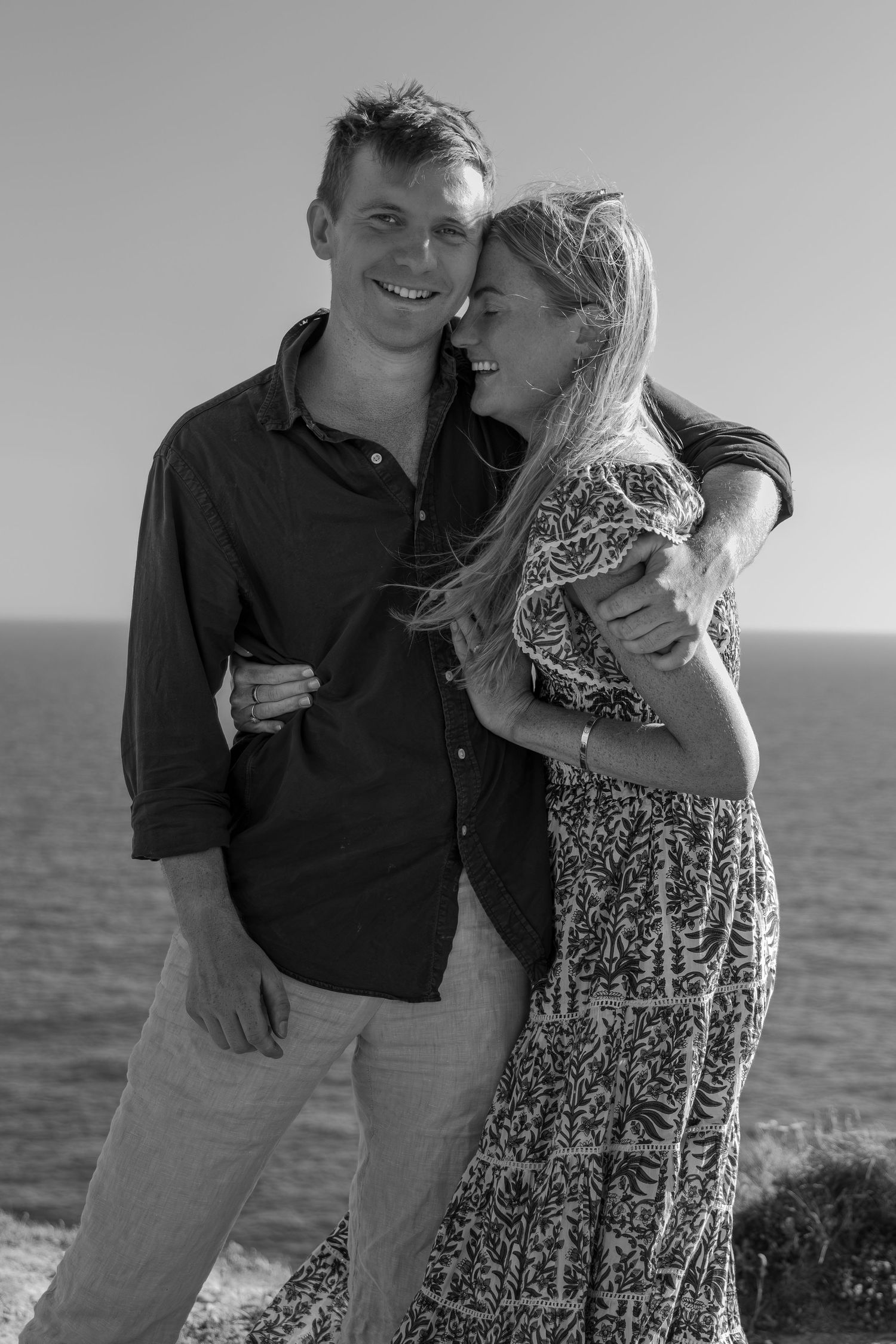 Black and white photo of a smiling couple embracing outdoors by the ocean on a sunny day.