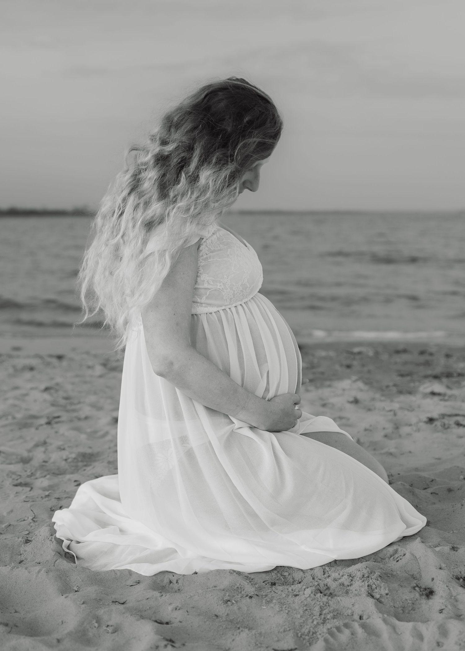 Pregnant woman in white dress sitting on beach sand, cradling baby bump, black and white maternity photo.