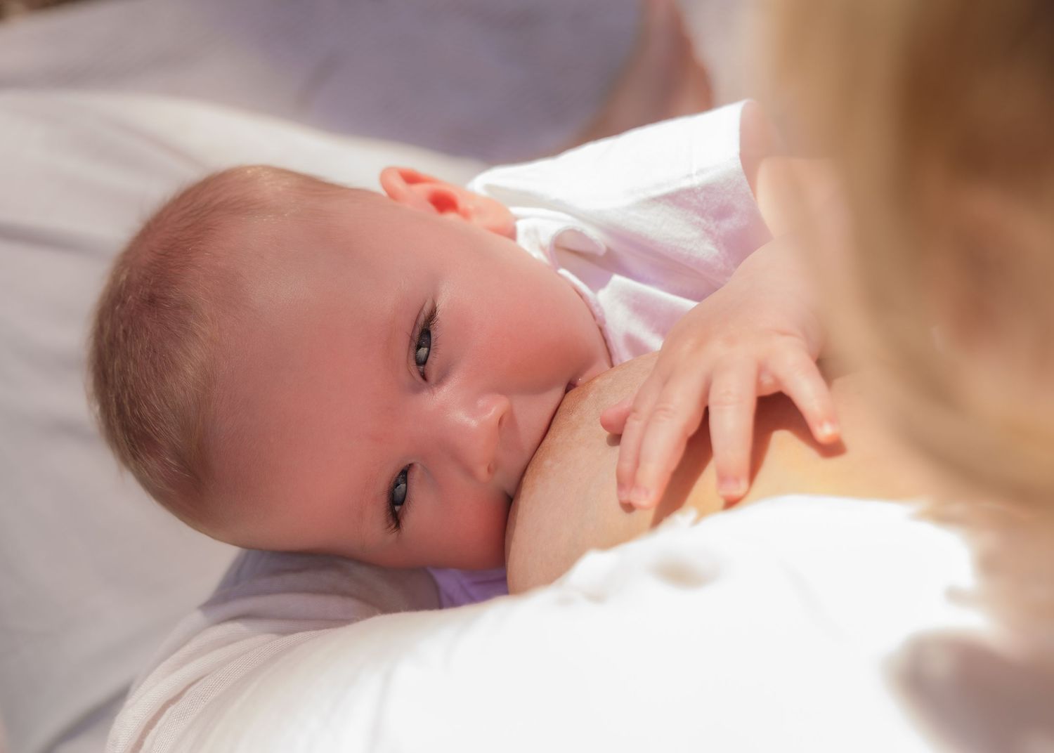 Newborn baby breastfeeding while gazing up at mother in soft natural light.