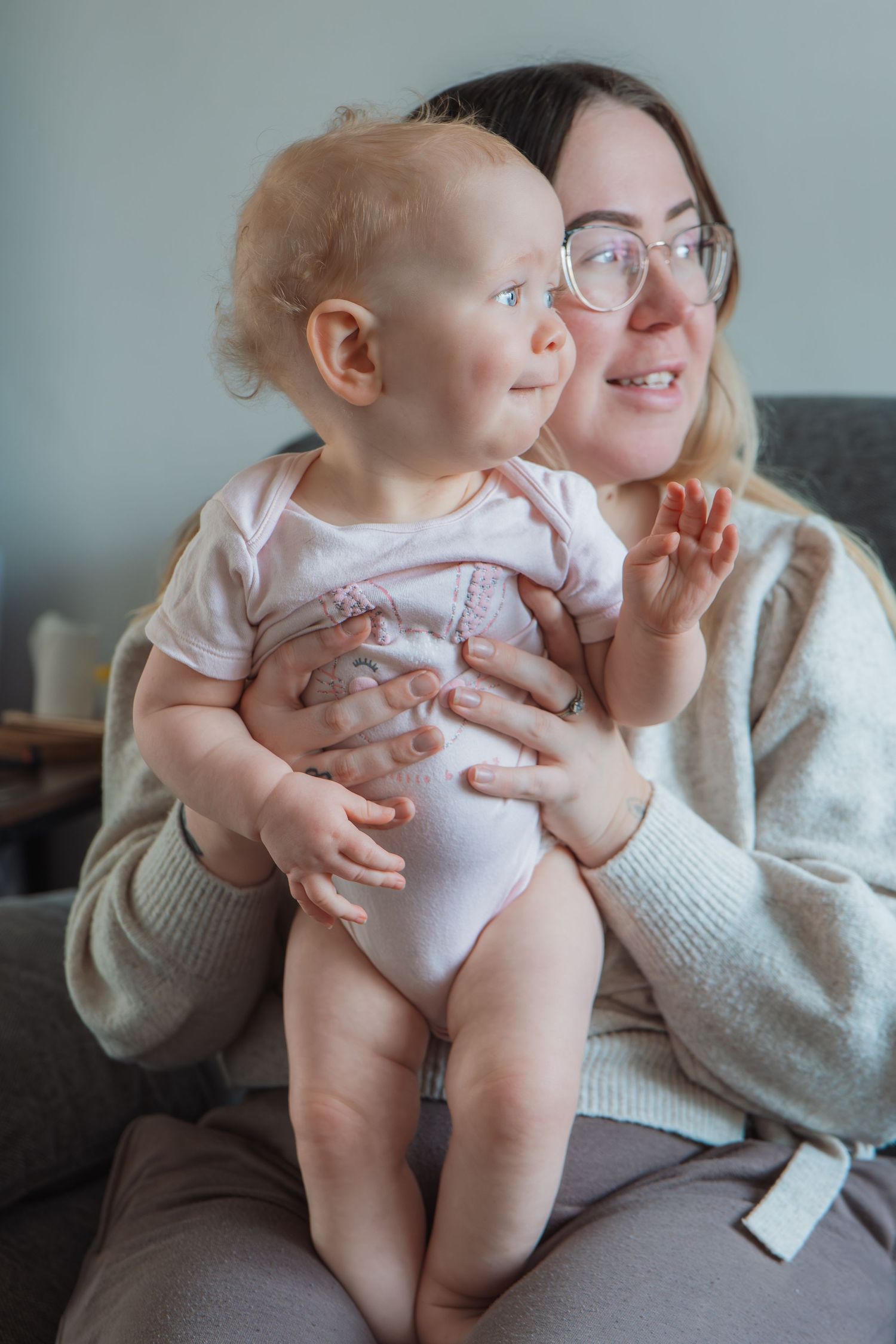 A smiling mother with glasses holds her happy baby in a pink onesie while sitting on a couch indoors.