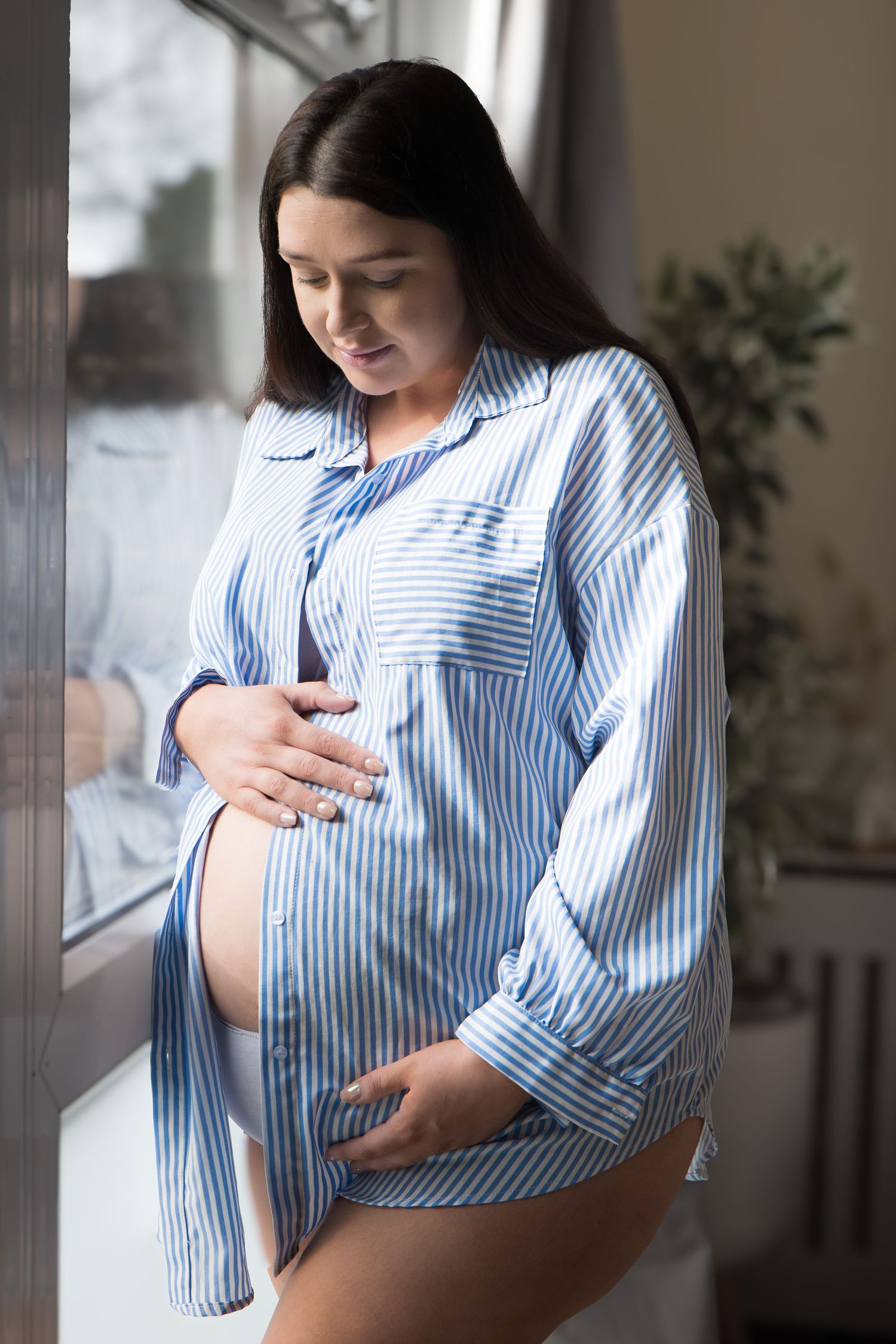 Pregnant woman in blue striped shirt standing by window cradling her bare baby bump.