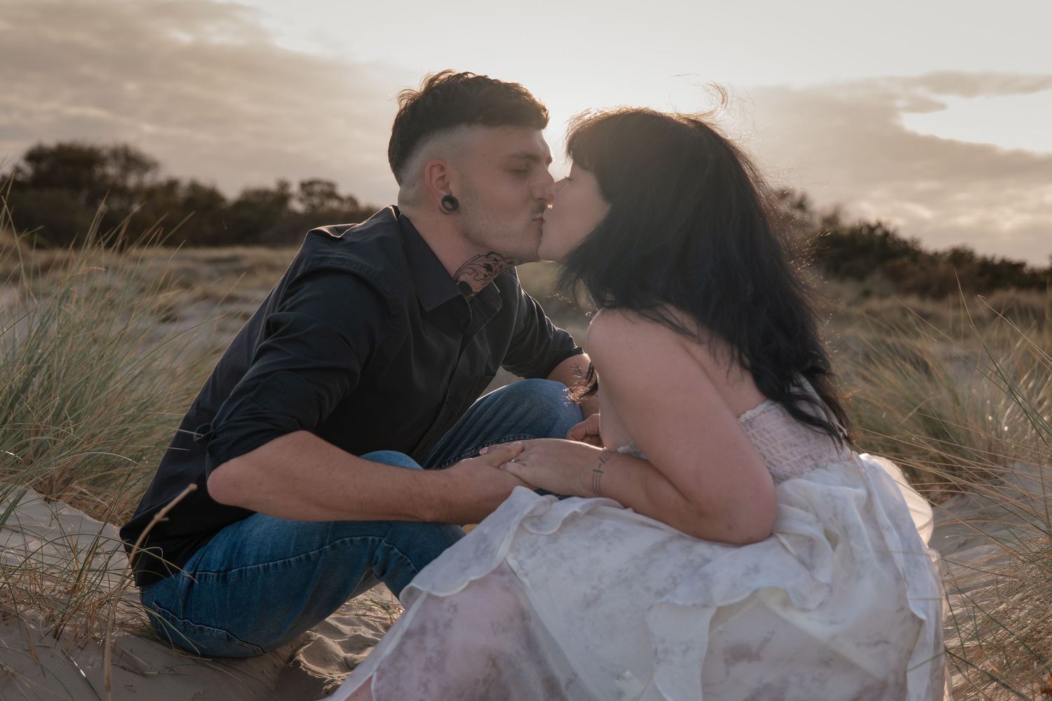 Couple sharing a romantic kiss while sitting on sandy dunes at sunset, woman in white dress, man in black shirt.