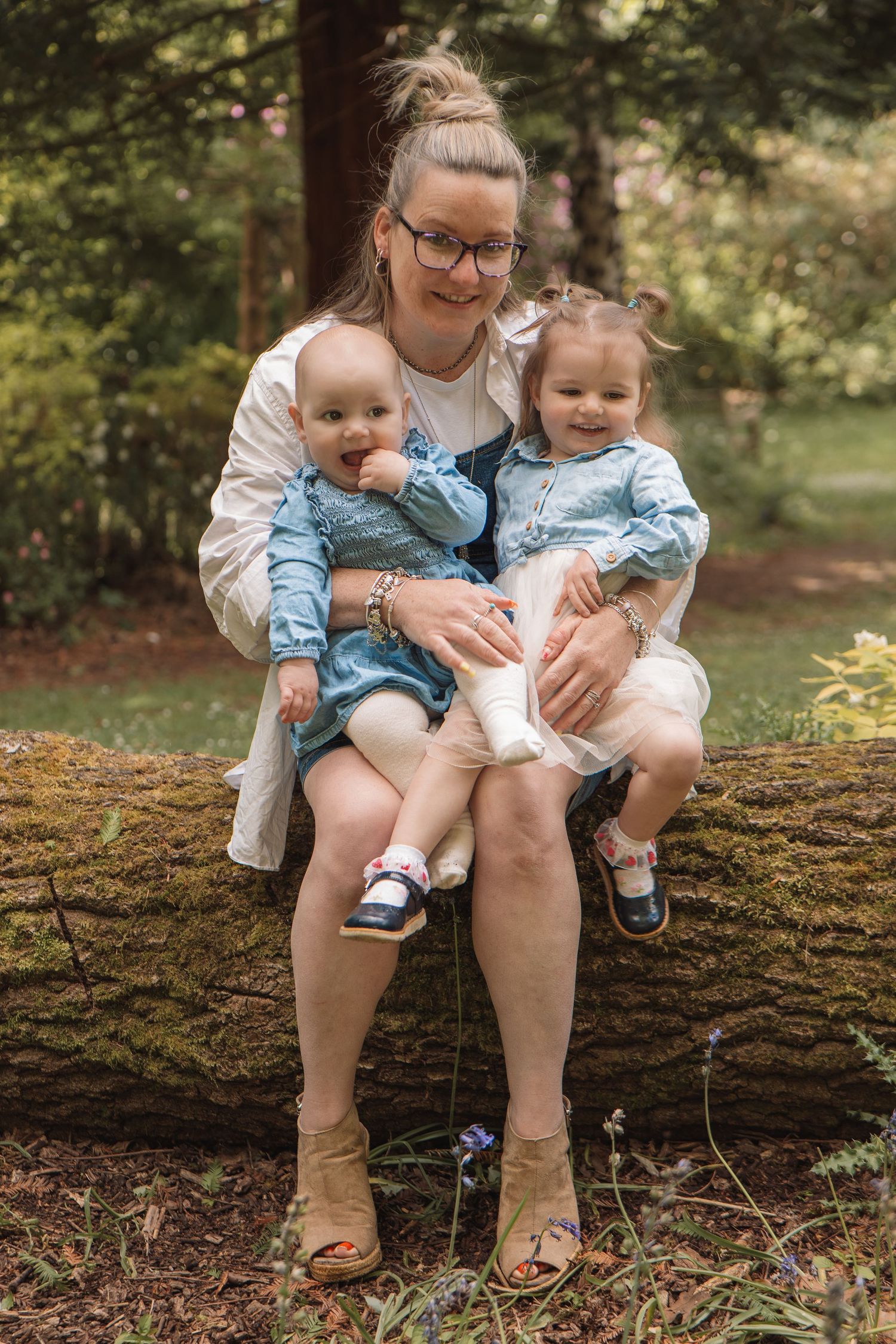A smiling mother sits on a mossy log in a forest holding two young daughters in matching blue dresses.