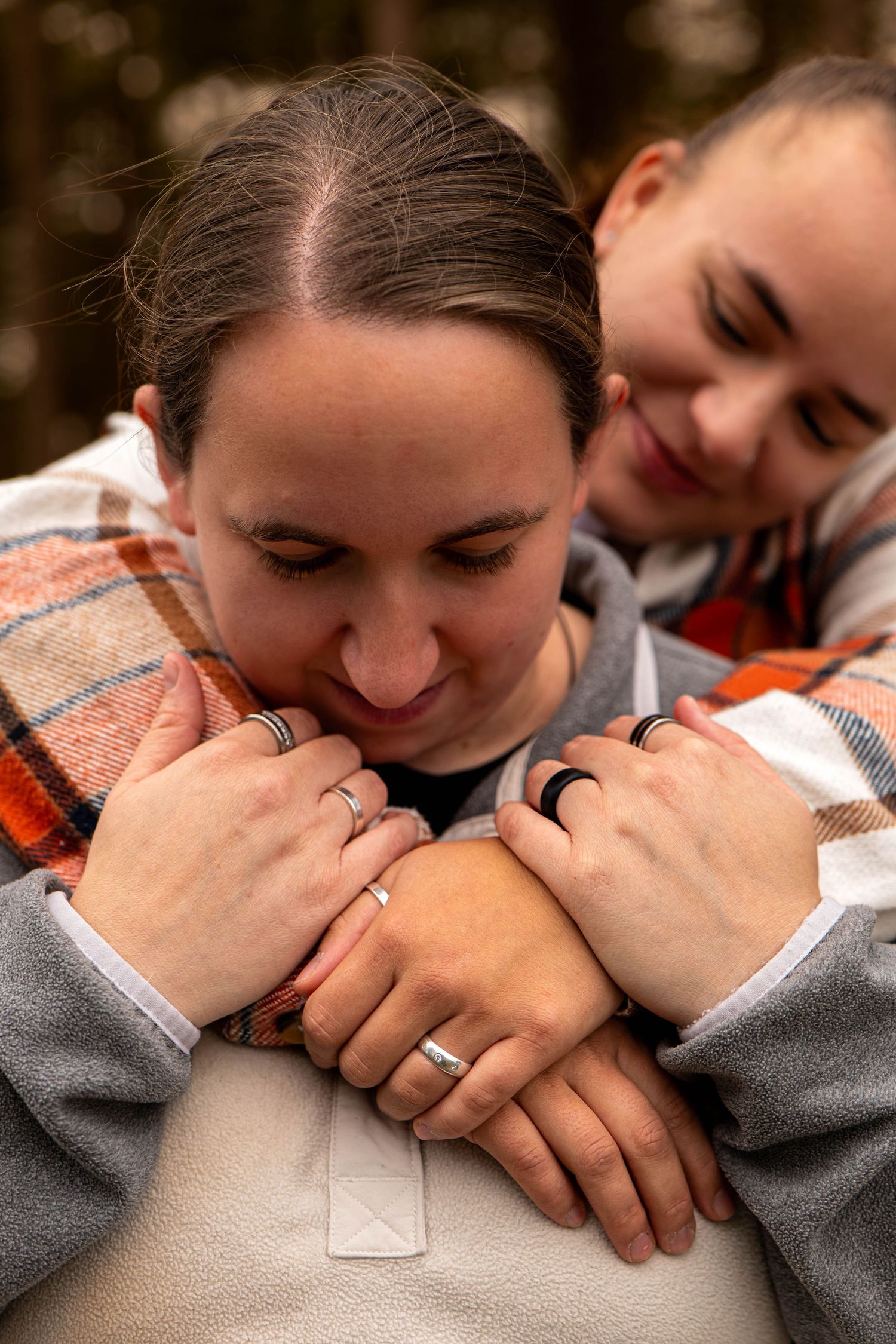 Couple embracing in forest with matching engagement rings visible on their intertwined hands.