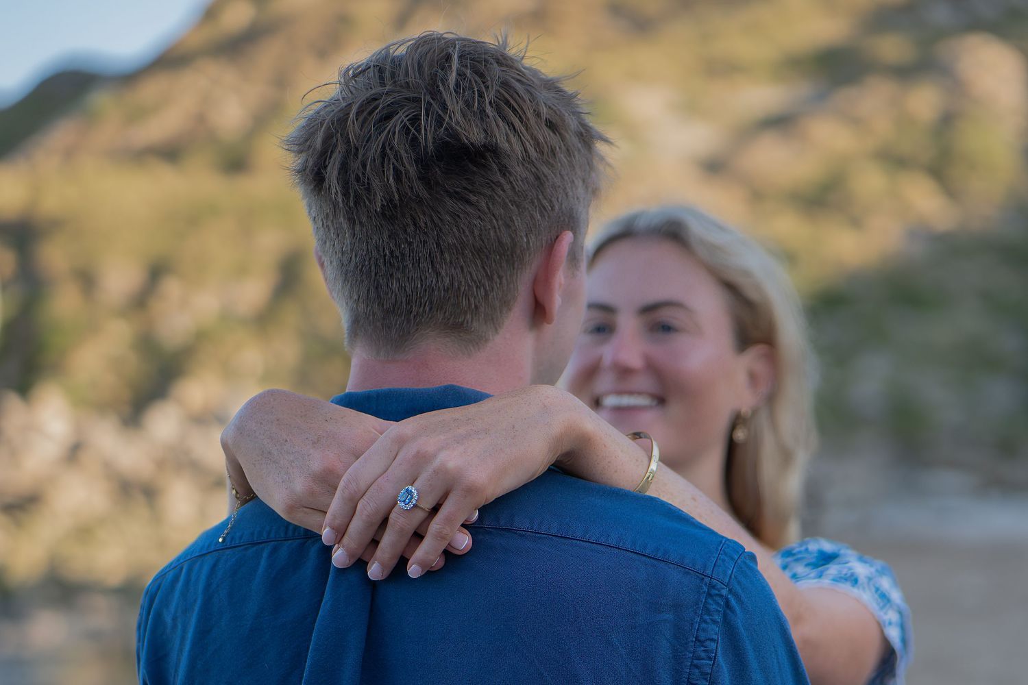 Couple embracing outdoors, woman smiling with engagement ring visible on hand resting on mans shoulder, mountain backdrop.