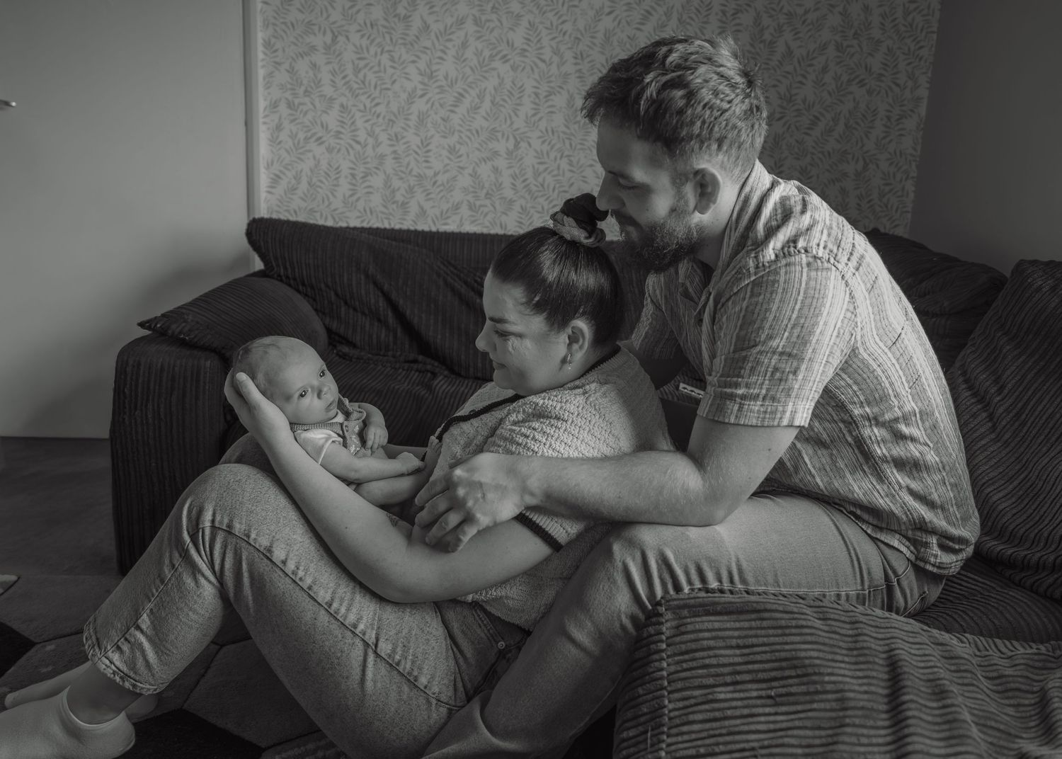 Black and white photo of parents sitting on floor admiring their newborn baby together at home.