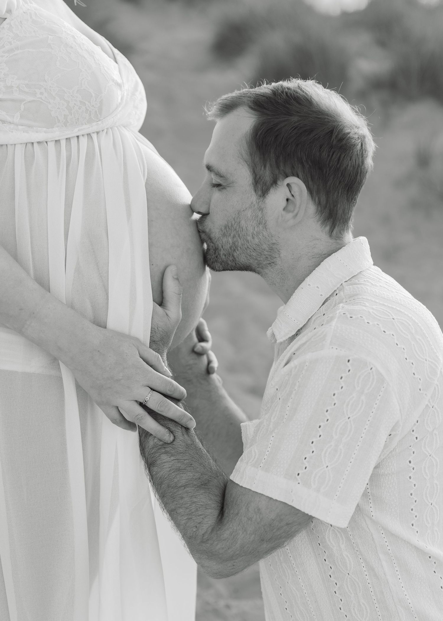 Black and white photo of a man kneeling and kissing a pregnant woman's belly during a maternity photoshoot outdoors.