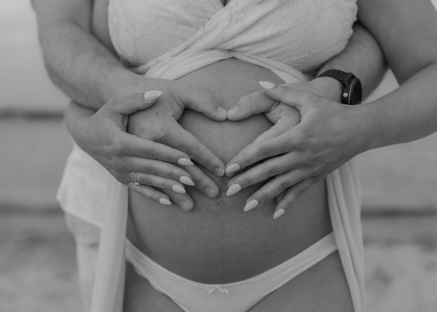 Black and white photo of pregnant couple forming heart shape with hands on baby bump at beach.