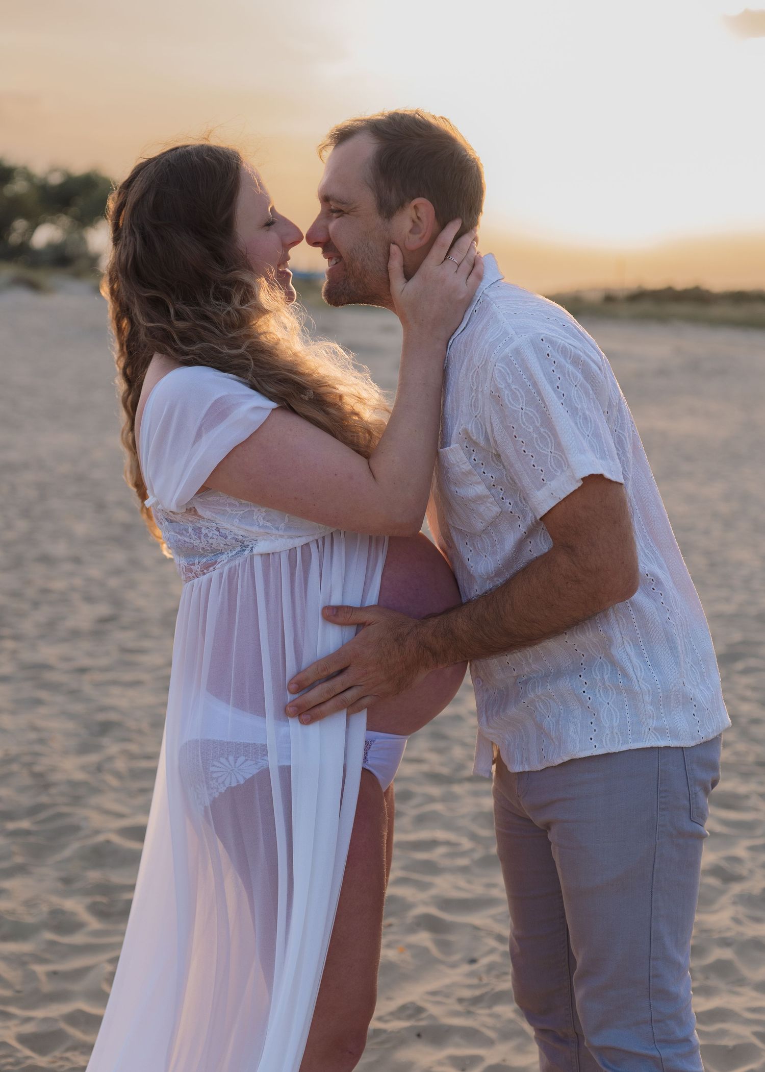 Pregnant couple sharing a romantic moment on a sandy beach at golden sunset, woman in white sheer maternity gown.