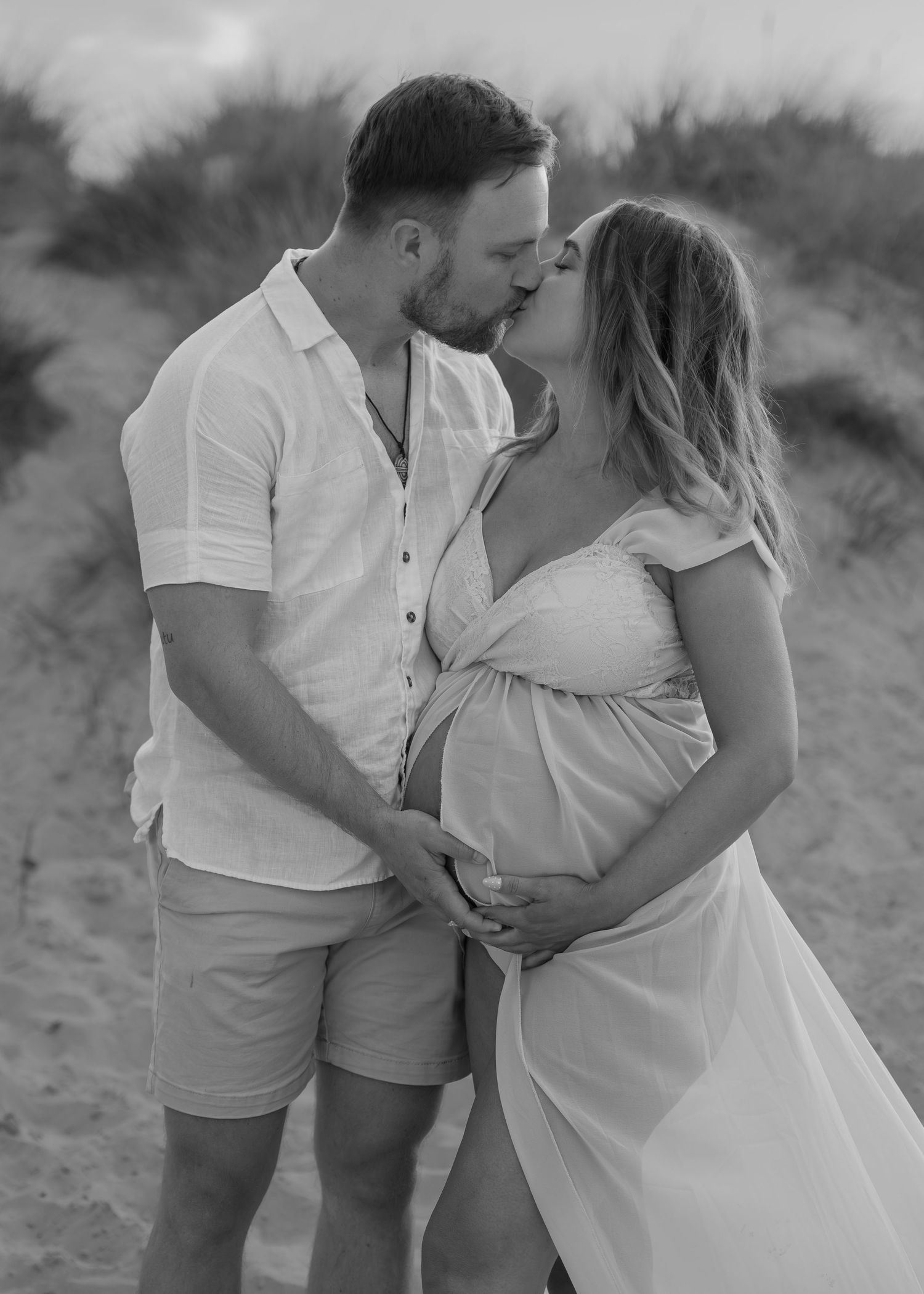 Black and white maternity photo of a couple kissing on a beach, cradling a pregnant belly, wearing white outfits.