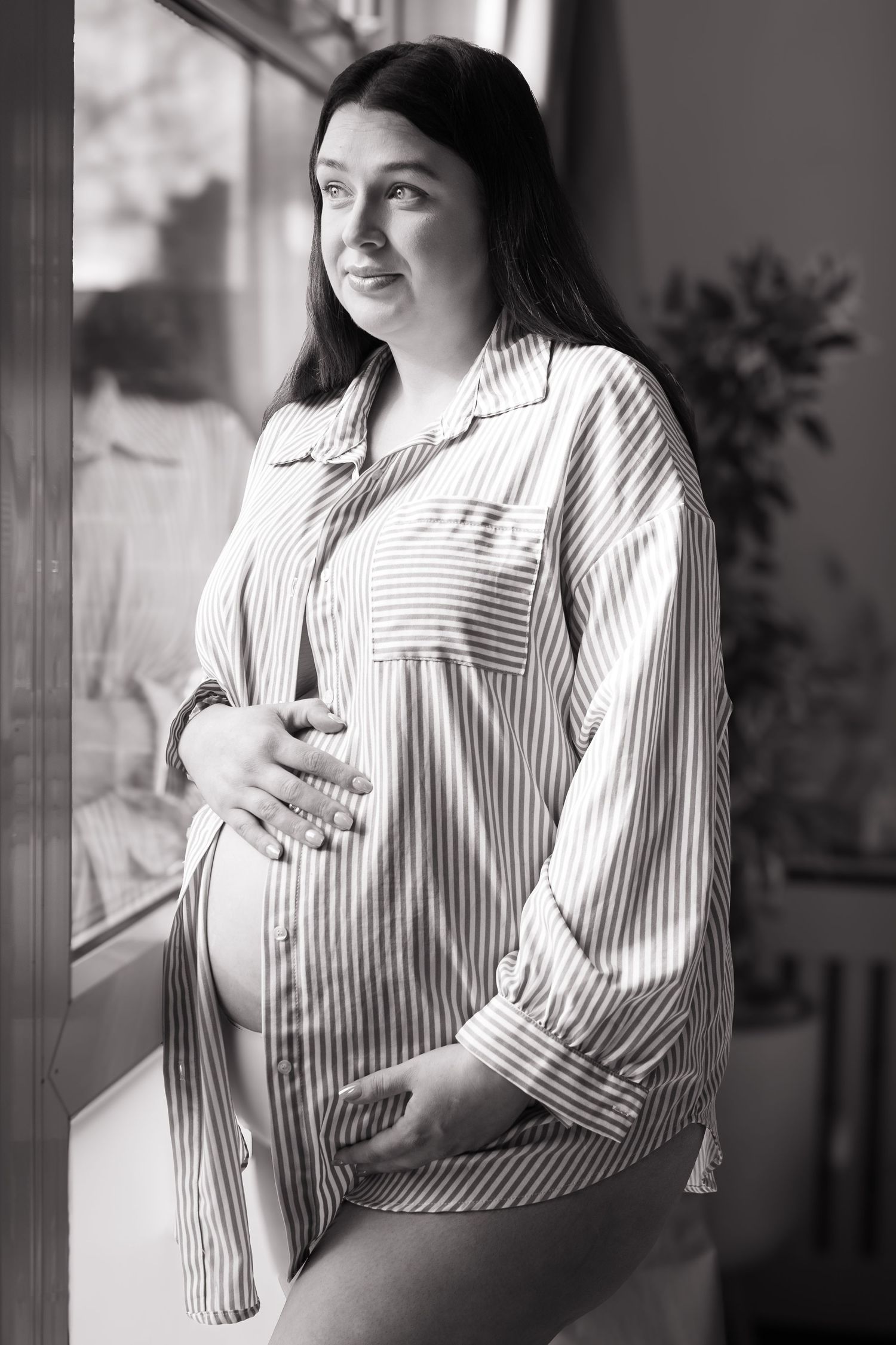 Black and white portrait of smiling pregnant woman in striped shirt standing by window indoors.