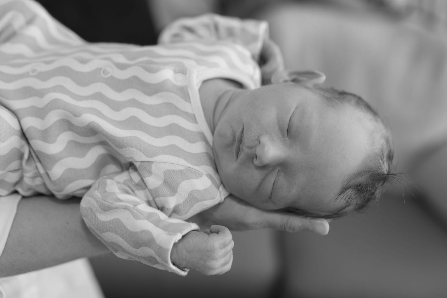 Black and white image of sleeping newborn baby in chevron onesie cradled in parent's arms.