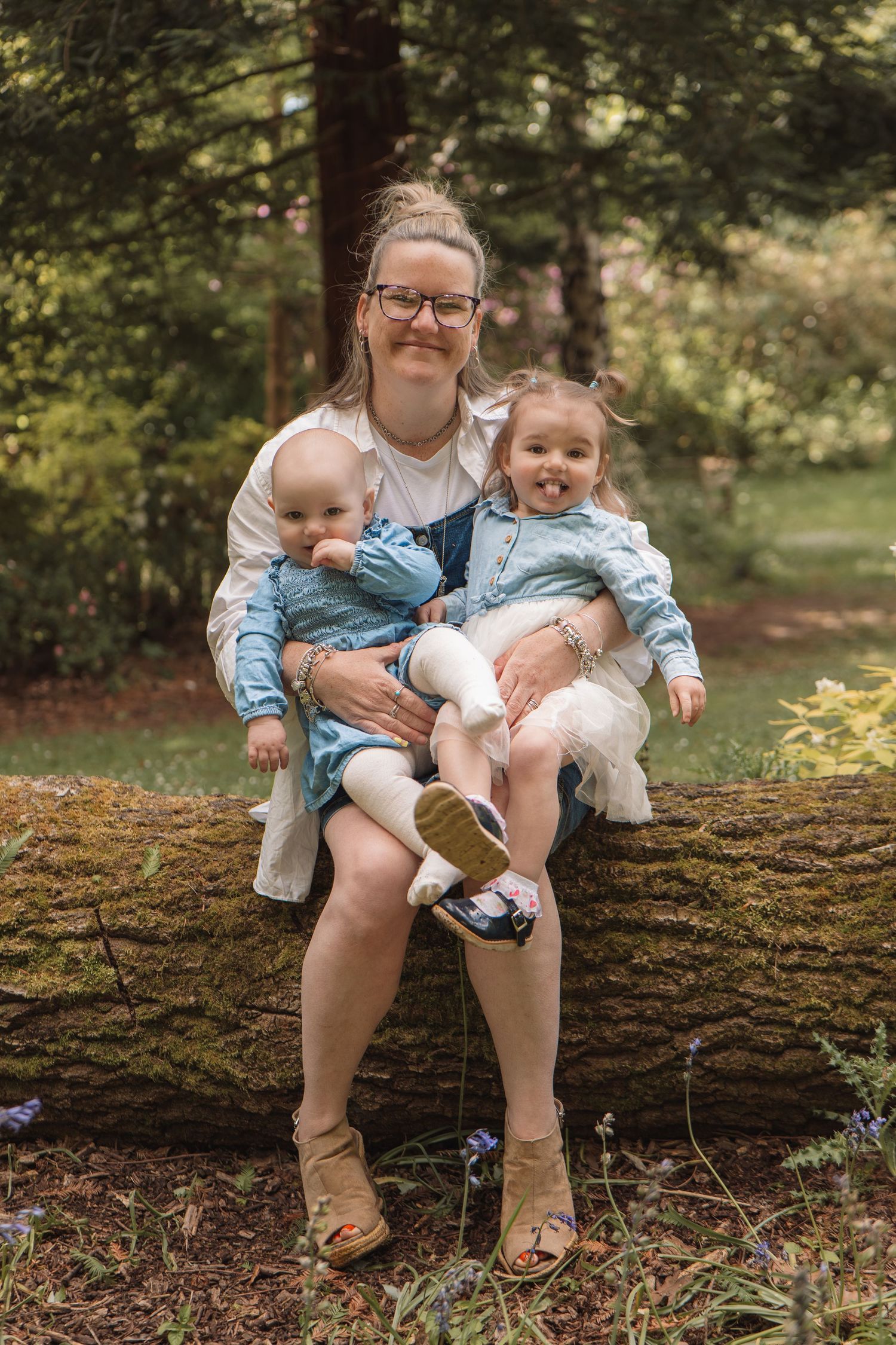 A woman sitting on a mossy log in a forest, smiling while holding two young girls in matching denim dresses.