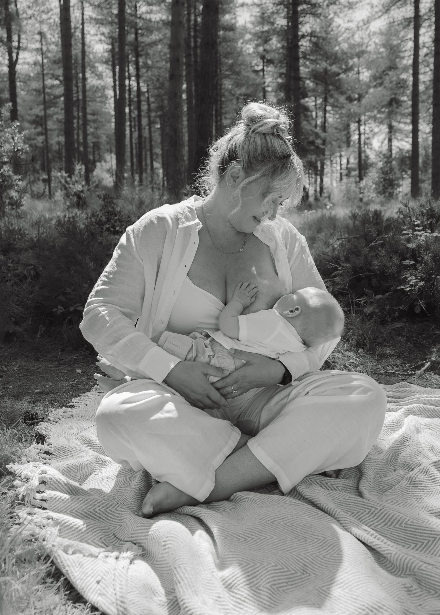 Black and white photo of a mother breastfeeding her newborn baby outdoors on a blanket in a forest.