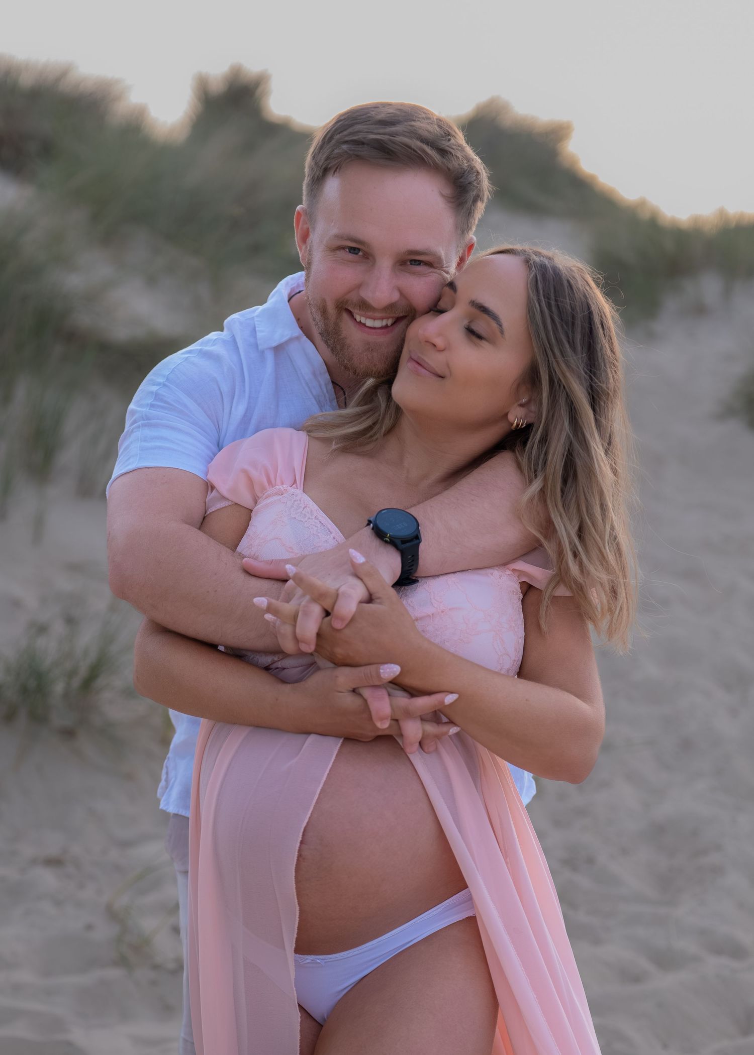 Couple embracing on sandy beach during maternity photoshoot, woman in pink dress showing baby bump at sunset.
