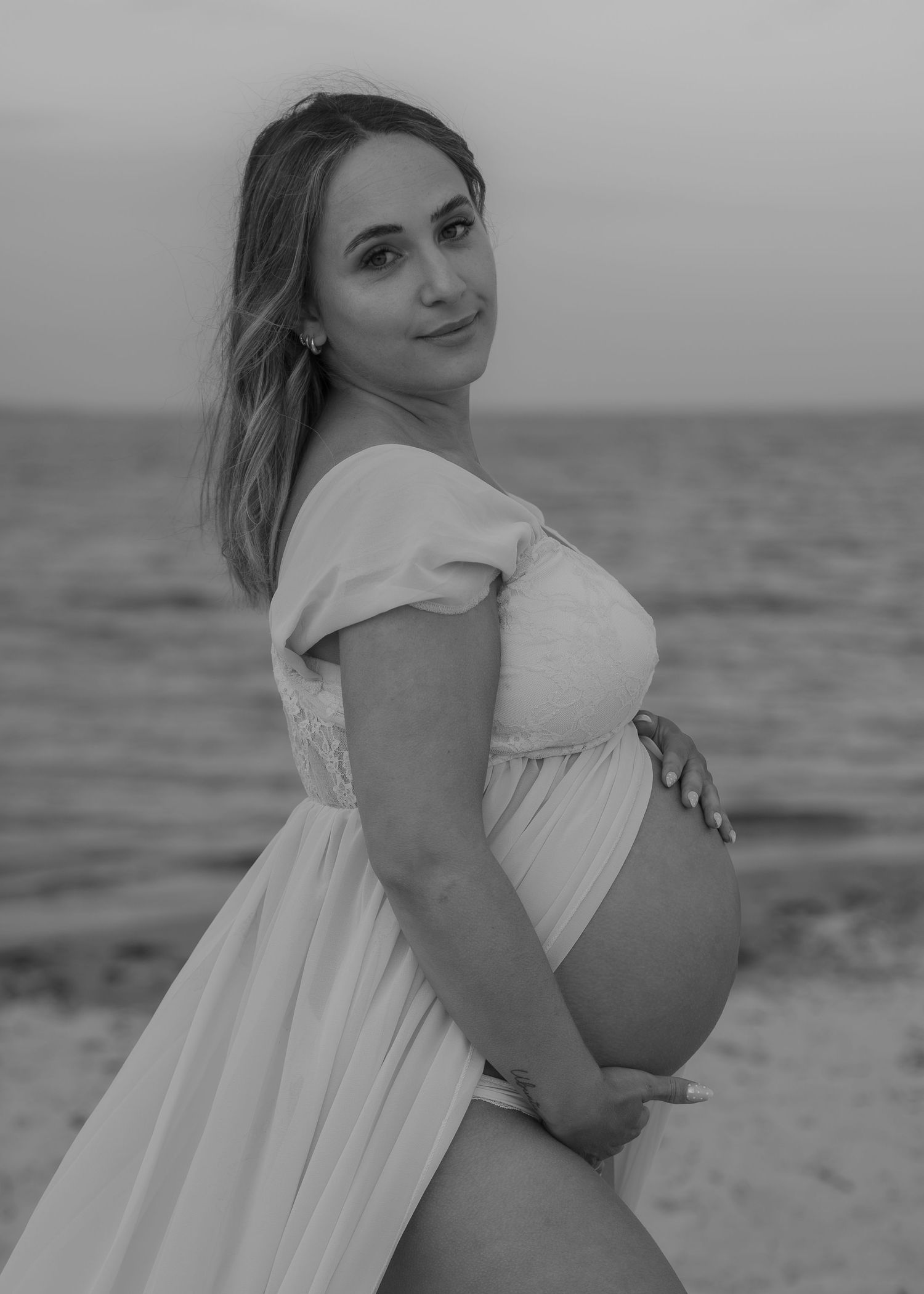 Black and white maternity photo of a pregnant woman in a white off-shoulder dress posing on a beach at sunset.