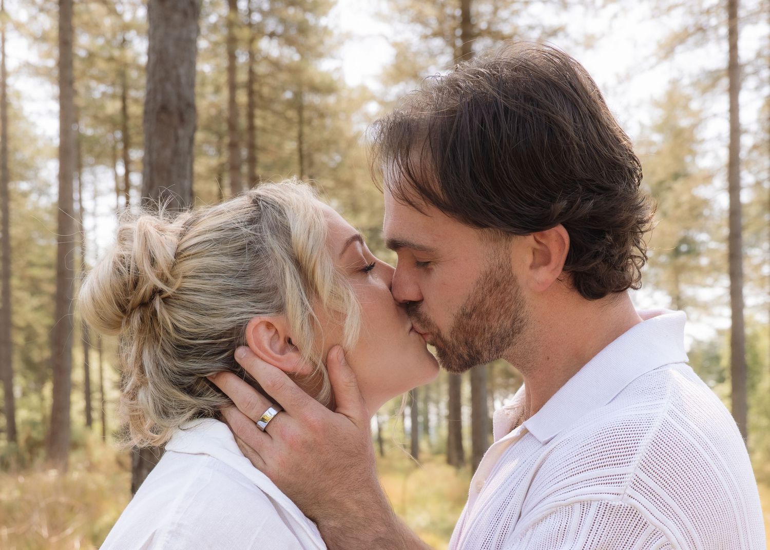 Romantic couple sharing a tender kiss outdoors among tall pine trees, woman with blonde hair wearing a ring.