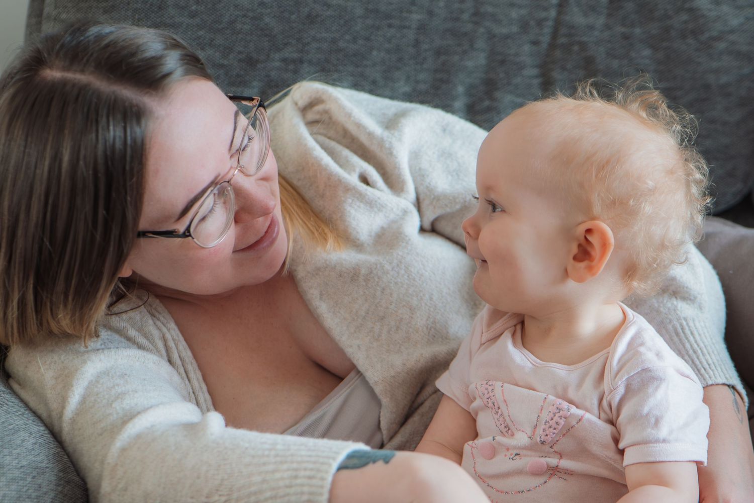 A young mother with glasses smiles at her happy baby while relaxing together on a cozy gray sofa.