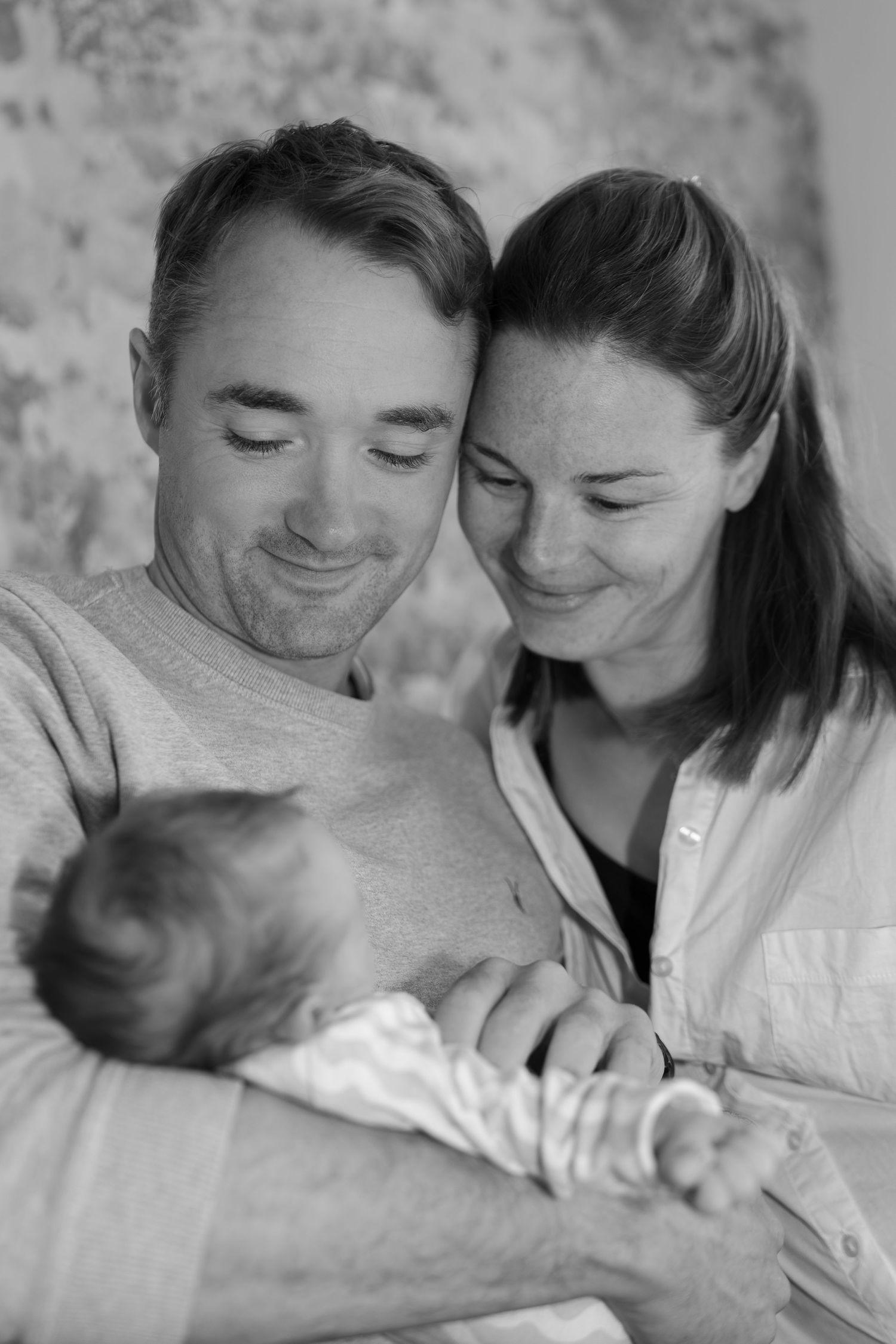 Black and white photo of smiling parents lovingly gazing down at their newborn baby.