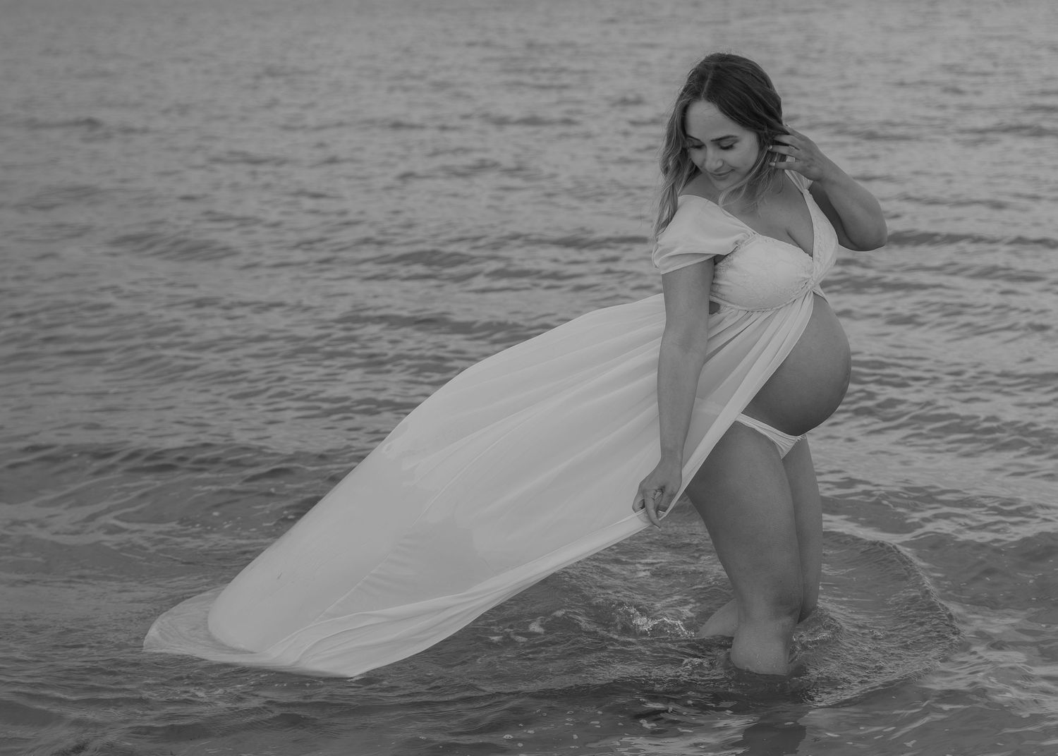 Pregnant woman in white flowing dress wading in ocean waves, elegant black and white maternity photography.