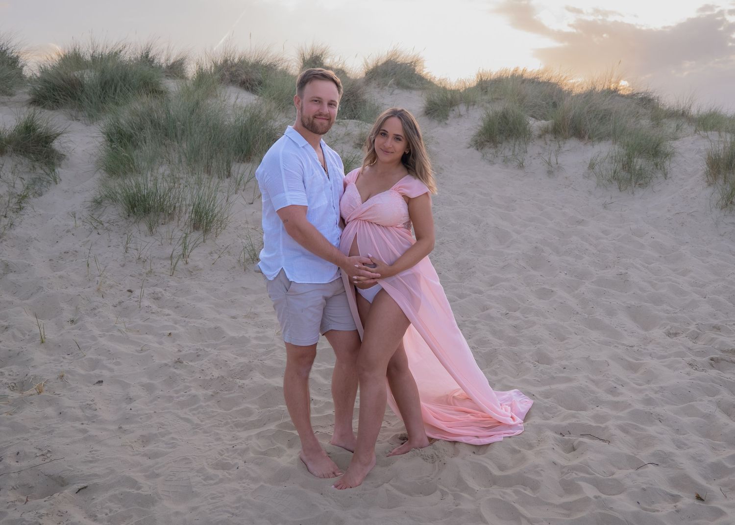 Pregnant couple posing on sandy beach at sunset, woman in pink maternity gown, man in white shirt and gray shorts.